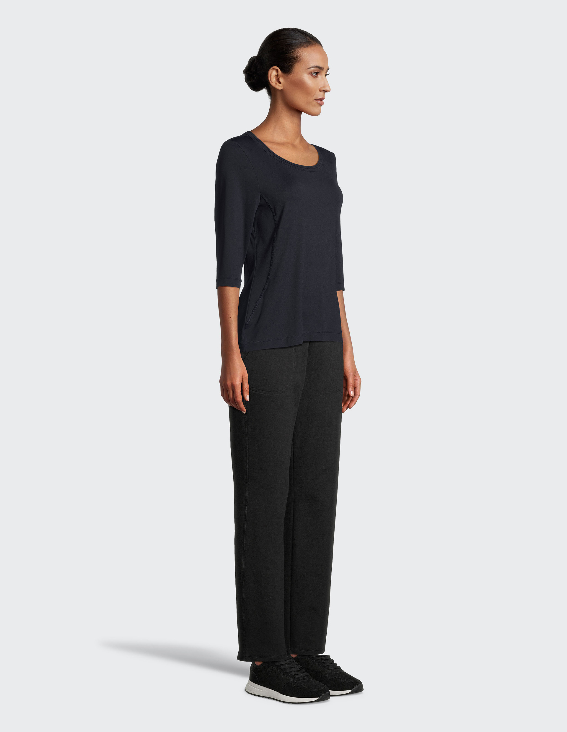 Frau in schwarzem Dreiviertelarmshirt und Hose, die seitlich steht, mit hochgestecktem Haar und schwarzen Sneakers auf grauem Hintergrund.