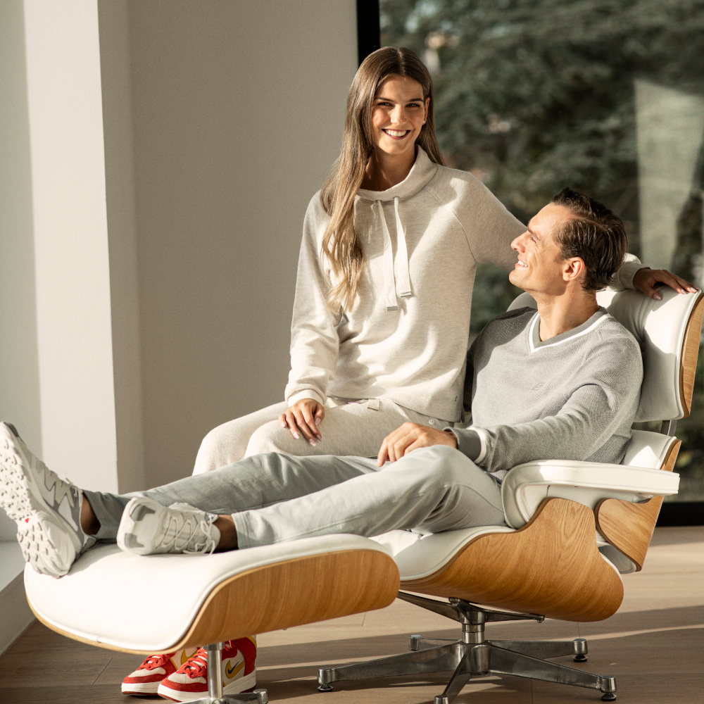 Mann trägt einen grauen Joghginganzug und sitzt in Stuhl. Frau sitzt auf Lehne des Stuhls und lächelt frontal in die Kamera.