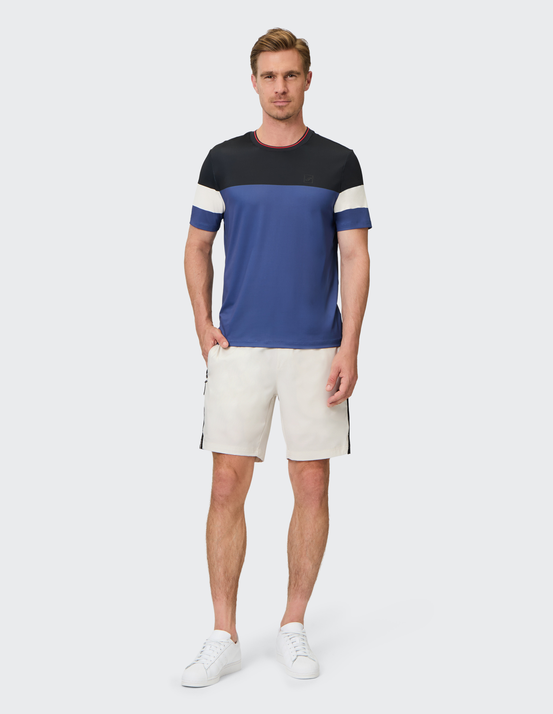 Ein Mann trägt ein blau-schwarz-weißes T-Shirt, weiße Shorts und weiße Sneakers vor neutralem Hintergrund.
