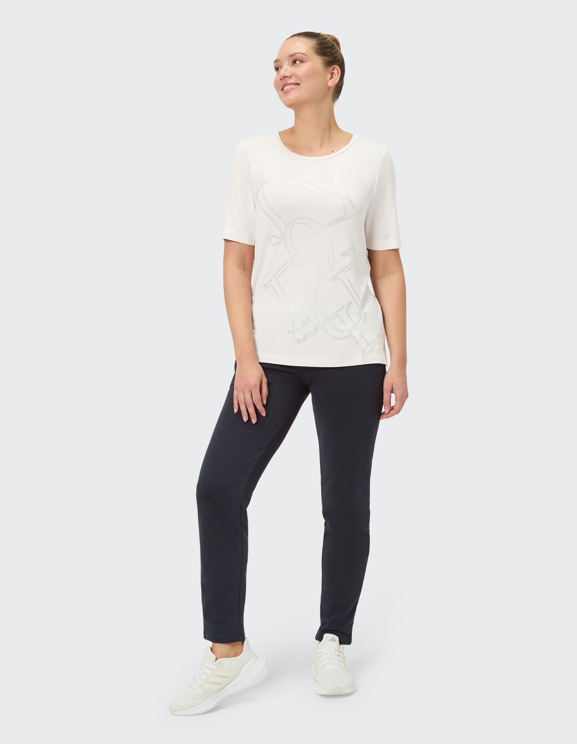 Person in weißem Oberteil mit dezentem Muster, dunkler Hose und weißen Sneakers, steht auf grauem Hintergrund.