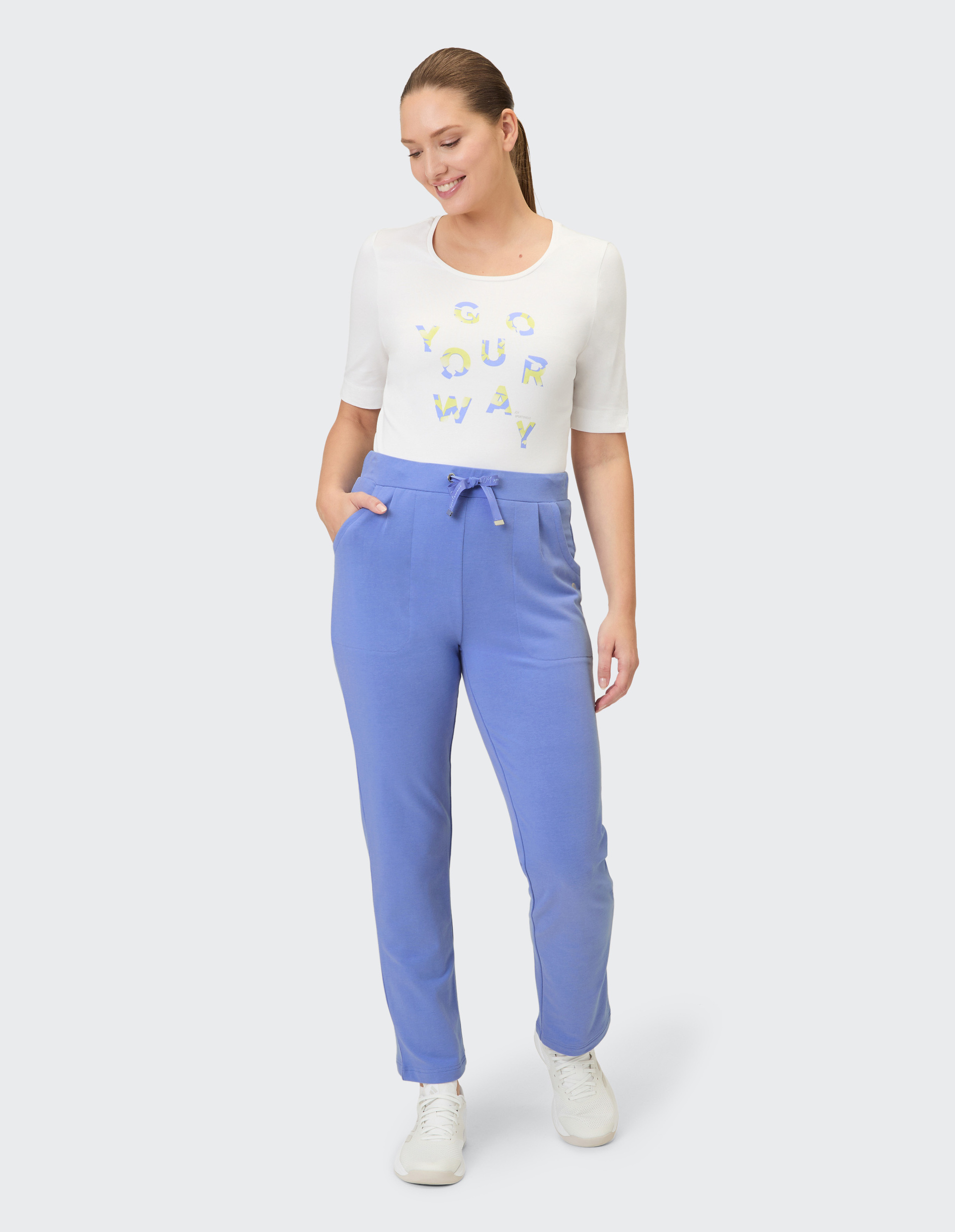 Eine Frau trägt eine weiße Kurzarmbluse mit bunter Schrift und blaue Stoffhose mit Tunnelzug.