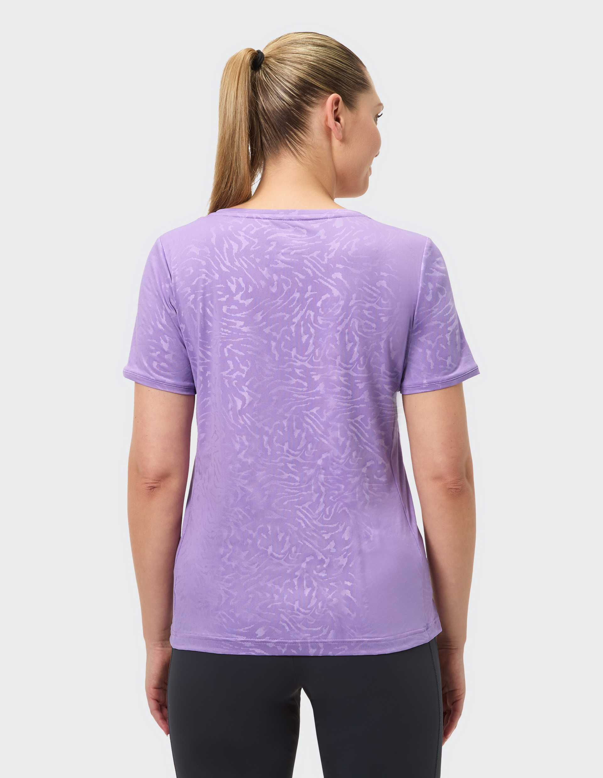 Frau mit Pferdeschwanz trägt ein lila T-Shirt mit subtilen Mustern, steht mit dem Rücken zur Kamera vor hellem Hintergrund.
