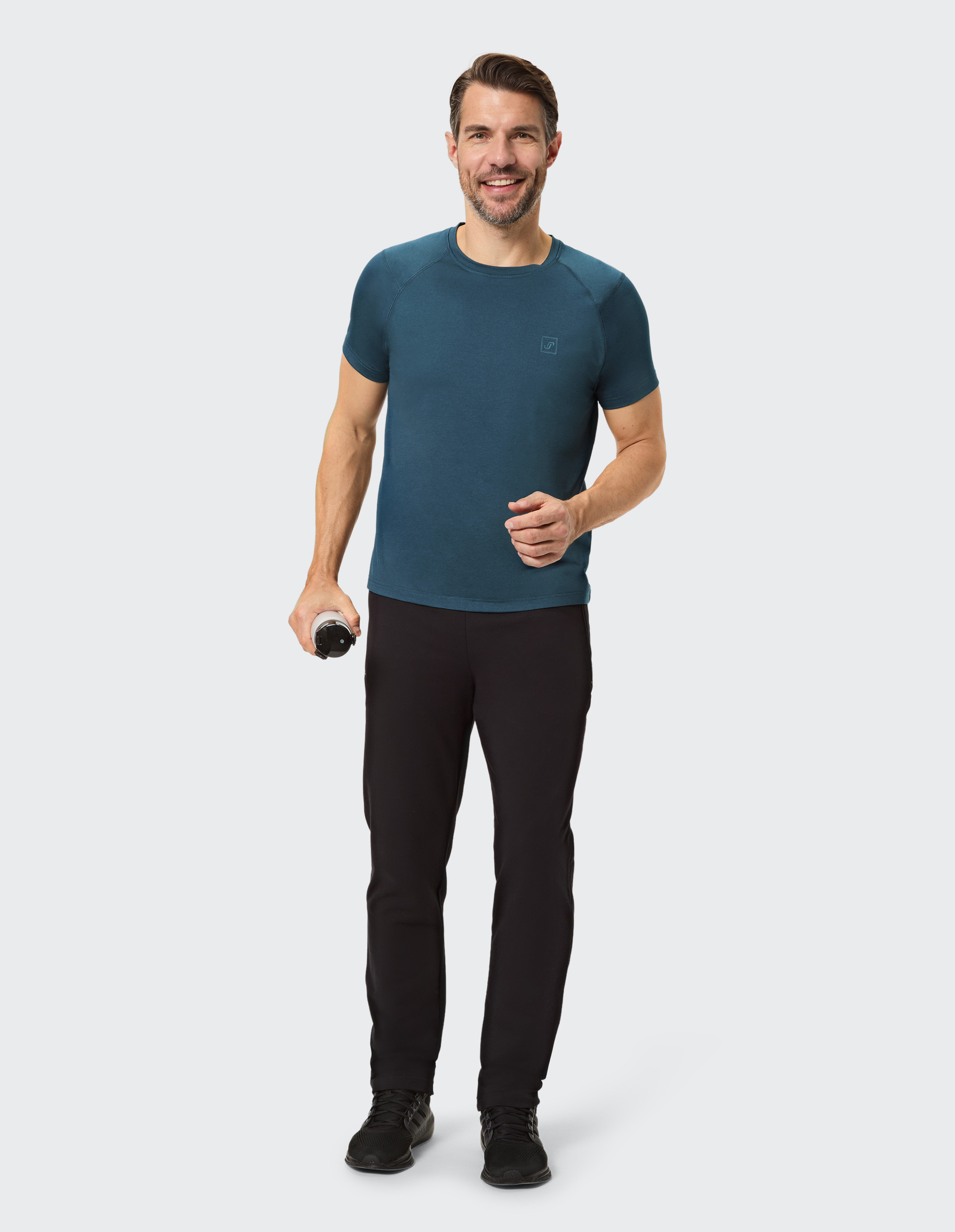 Ein Mann in einem dunkelblauen T-Shirt und schwarzen Hosen steht frontal vor einem hellen Hintergrund, beide Arme entspannt.