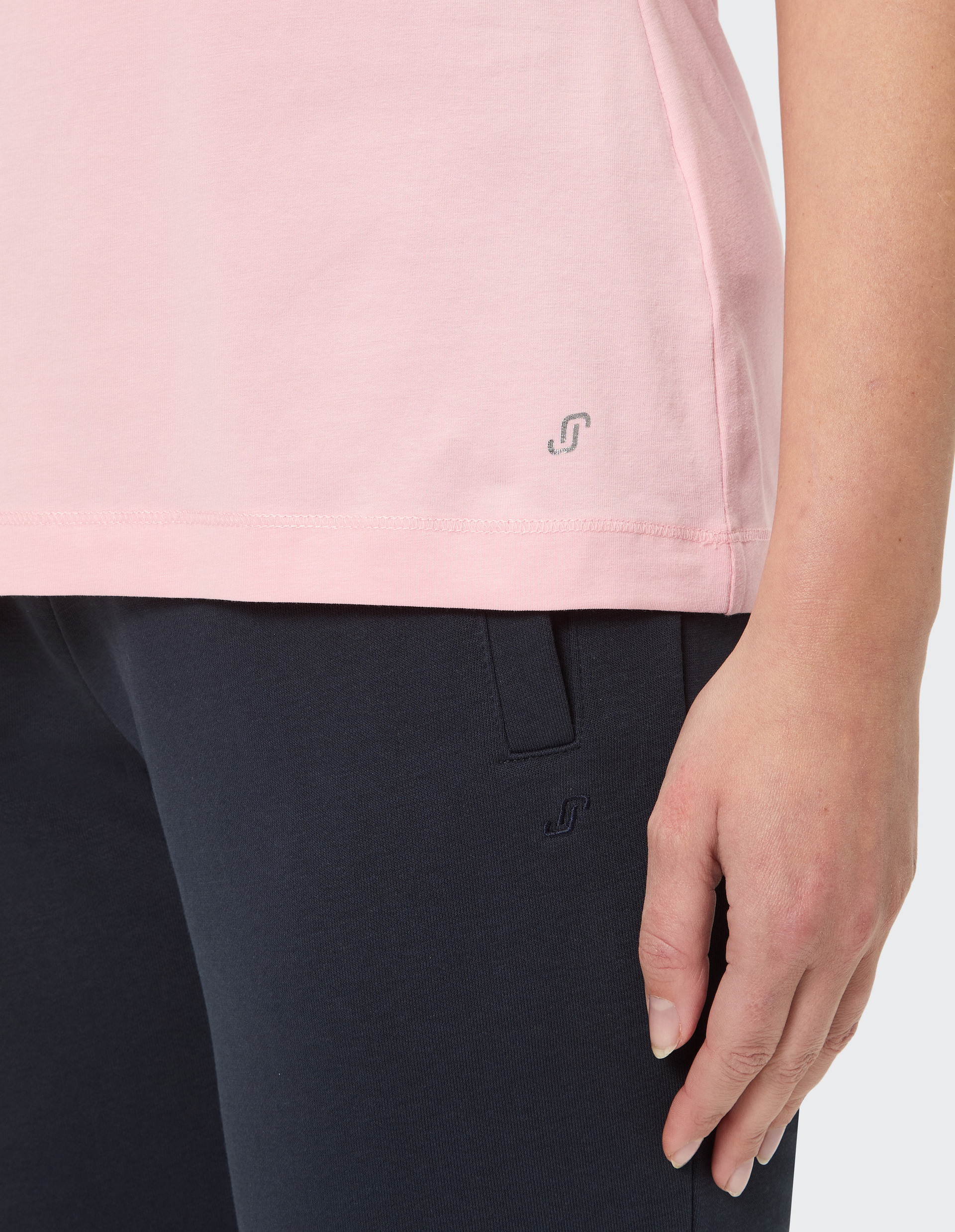 Unterer Teil eines rosa T-Shirts mit Logo und blauer Hose, rechte Hand einer Frau entspannt an der Seite hängend.