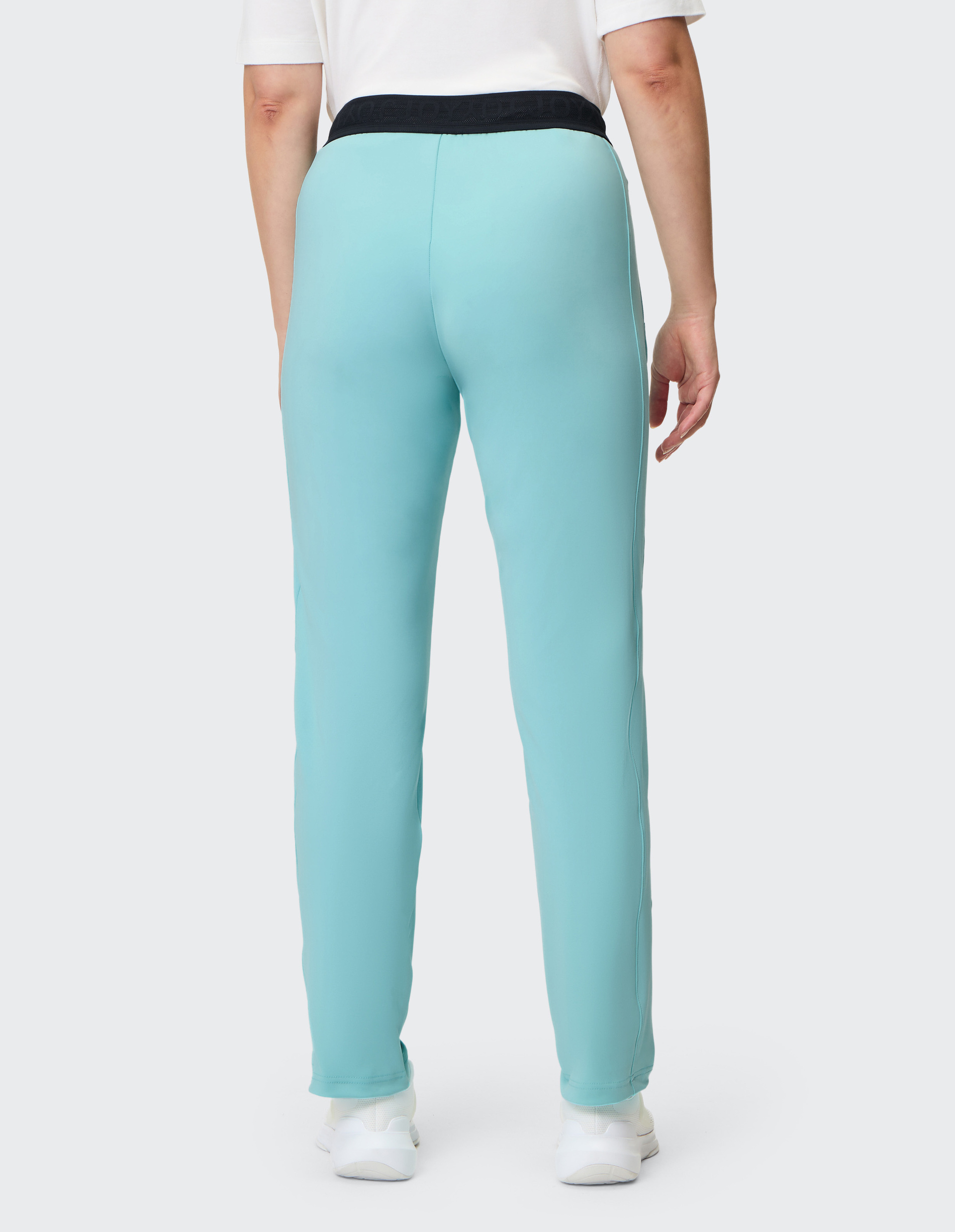 Person von hinten in türkisfarbener Hose mit schwarzem Bund, weißem T-Shirt und weißen Schuhen auf neutralem Hintergrund.