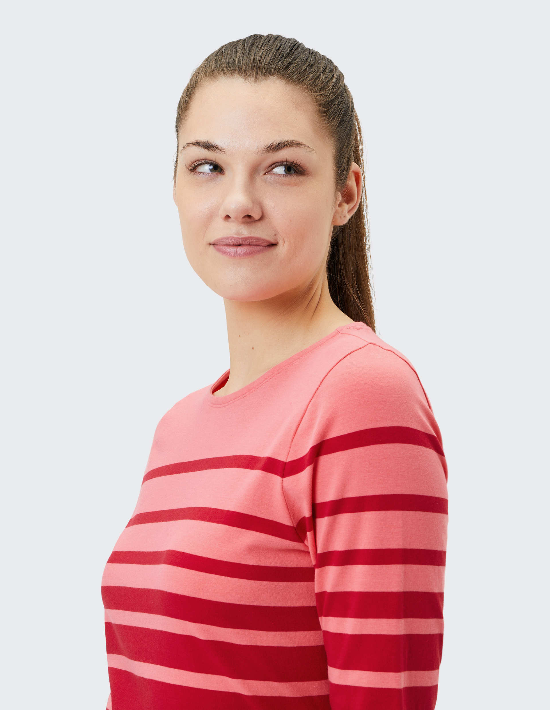 Frau mit langen Haaren trägt rosa-rot gestreiftes Oberteil, lächelt leicht und schaut nach links, vor grauem Hintergrund.