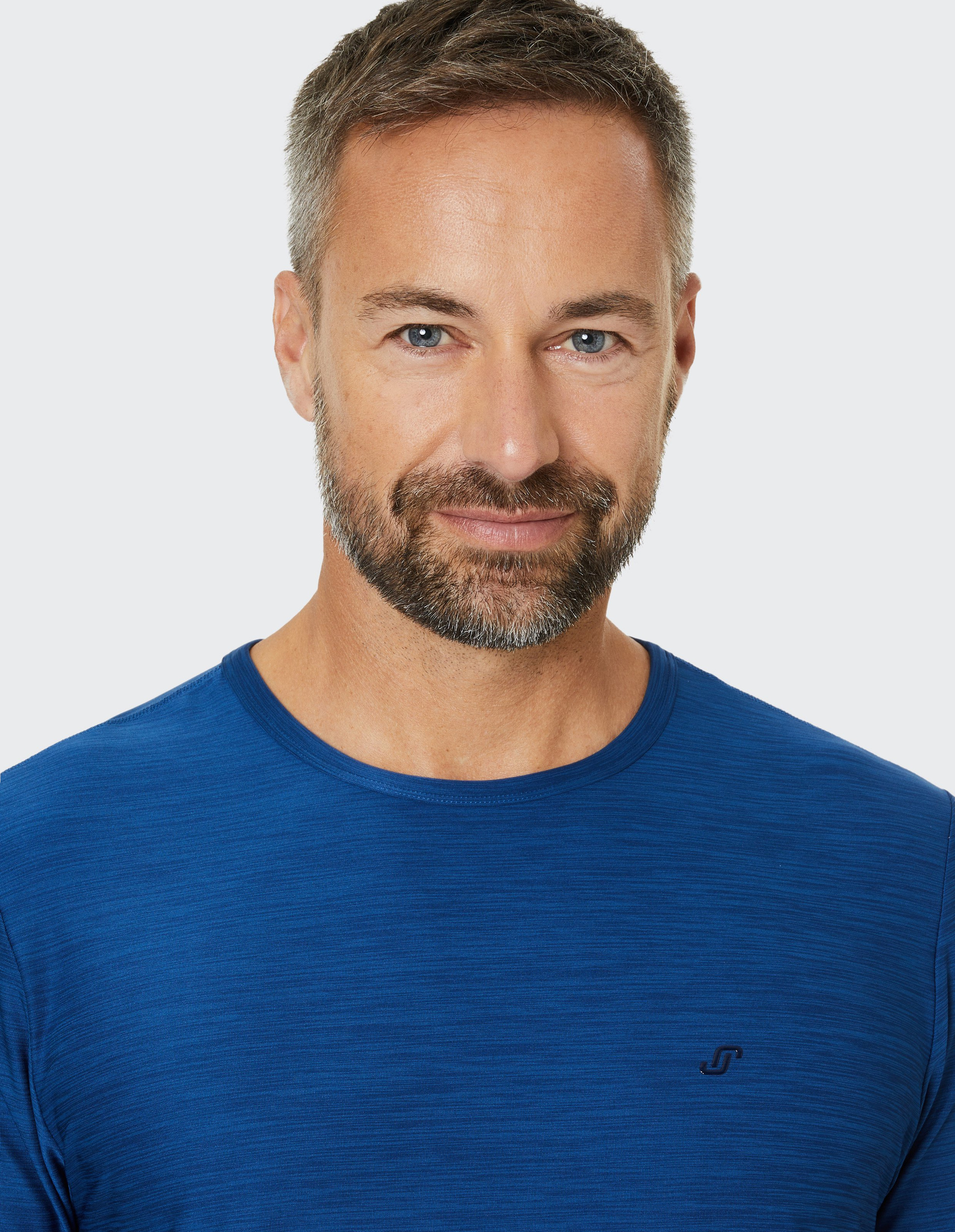 Ein Mann mit Bart und blauen Augen lächelt leicht, trägt ein blaues T-Shirt und hat kurzes, graues Haar, vor grauem Hintergrund.
