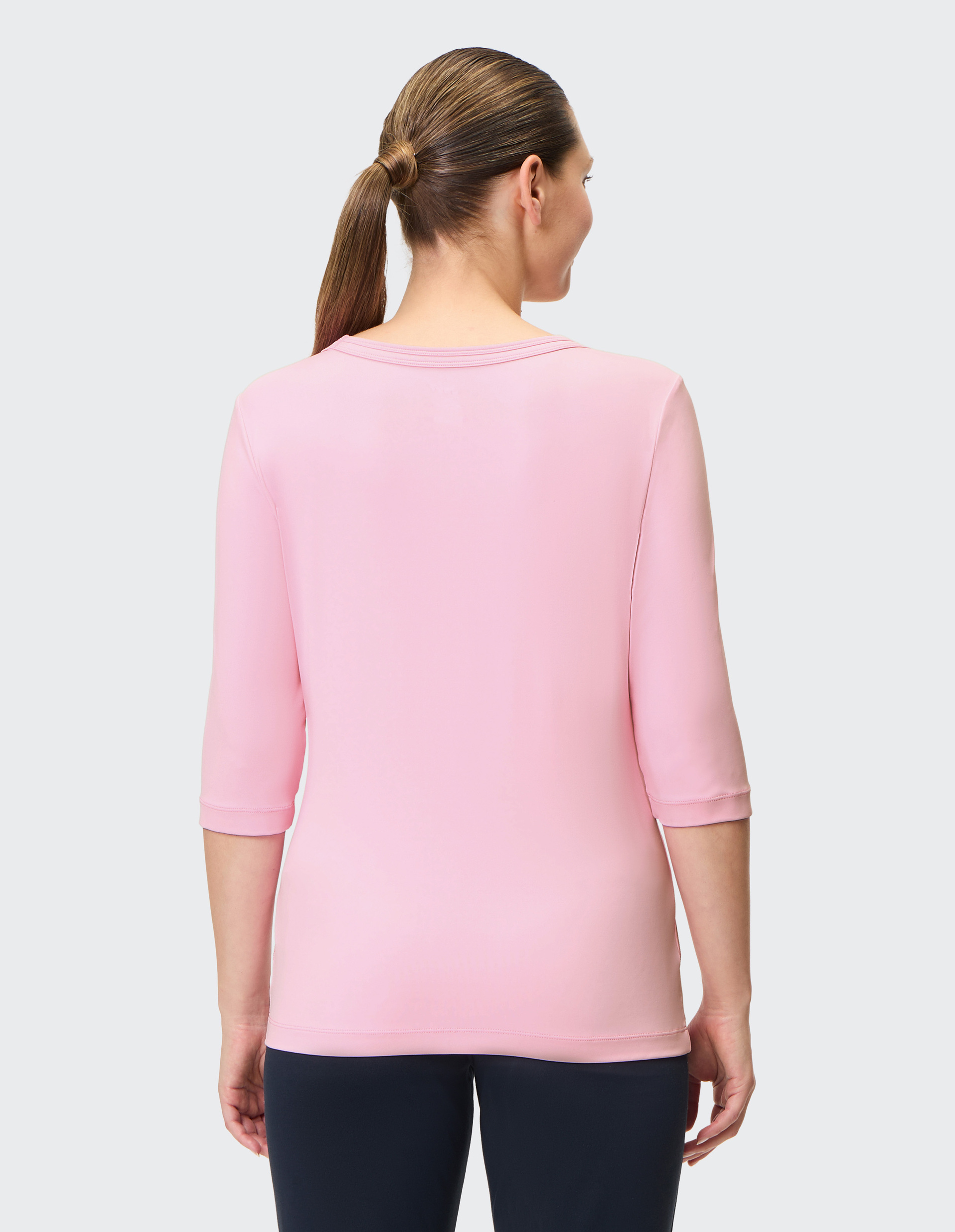 Eine Person mit langen Haaren, die einen Pferdeschwanz trägt, steht in einem rosa Shirt mit Dreiviertelärmeln und blickt nach rechts.