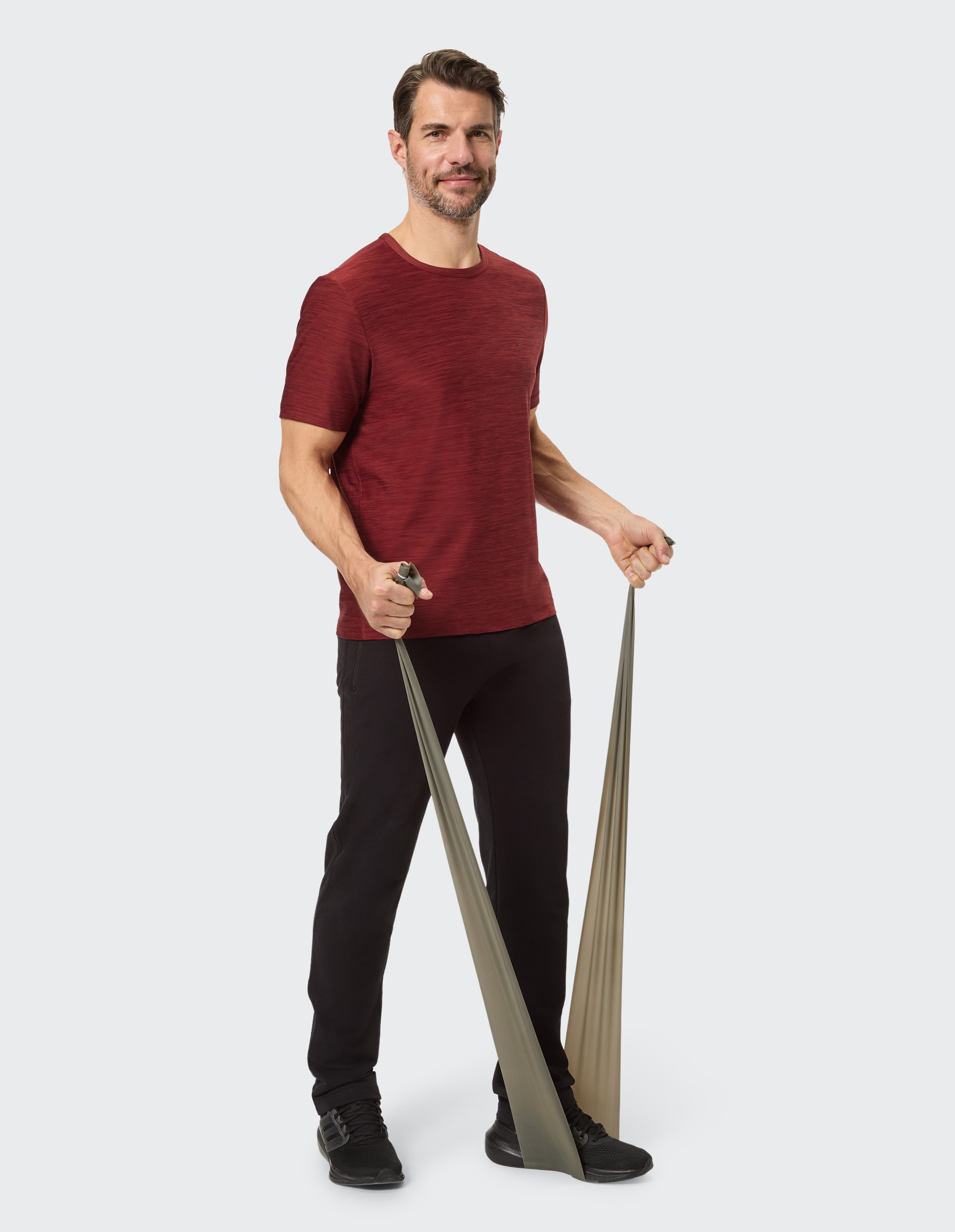 Ein lächelnder Mann in einem bordeauxroten T-Shirt und schwarzen Hosen steht freundlich posierend vor einem hellen Hintergrund.