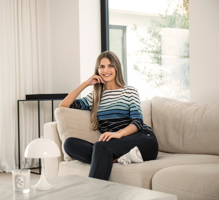 Auf dem Sofa sitzende Frau in dunkelblauer Hose und weiß, hellblau, blau und dunkelblau gestreiftem 3/4-Arm-Shirt der Marke JOY sportswear.