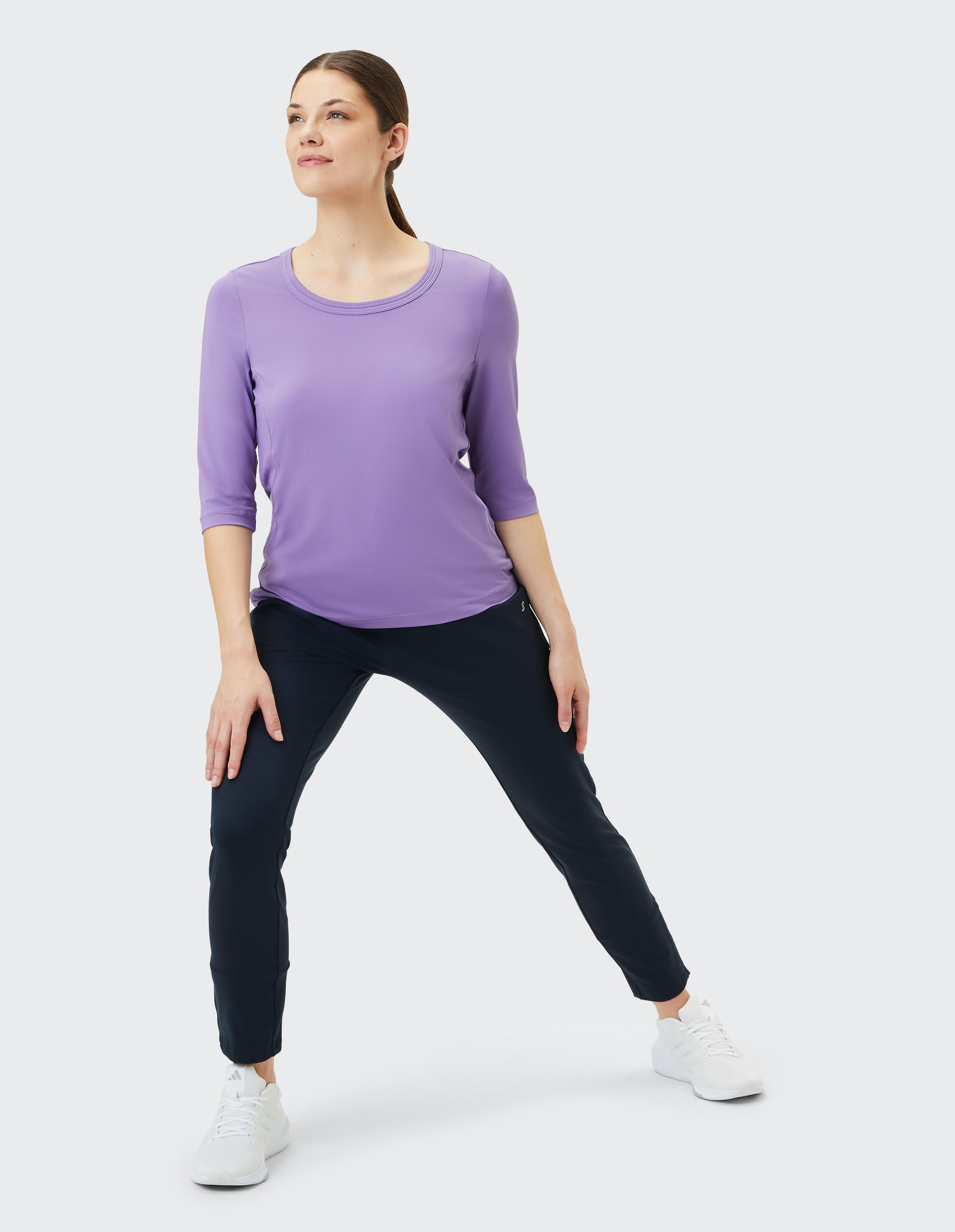 Frau in sportlicher Pose trägt ein lila Oberteil, dunkelblaue Hose und weiße Turnschuhe auf hellem Hintergrund.
