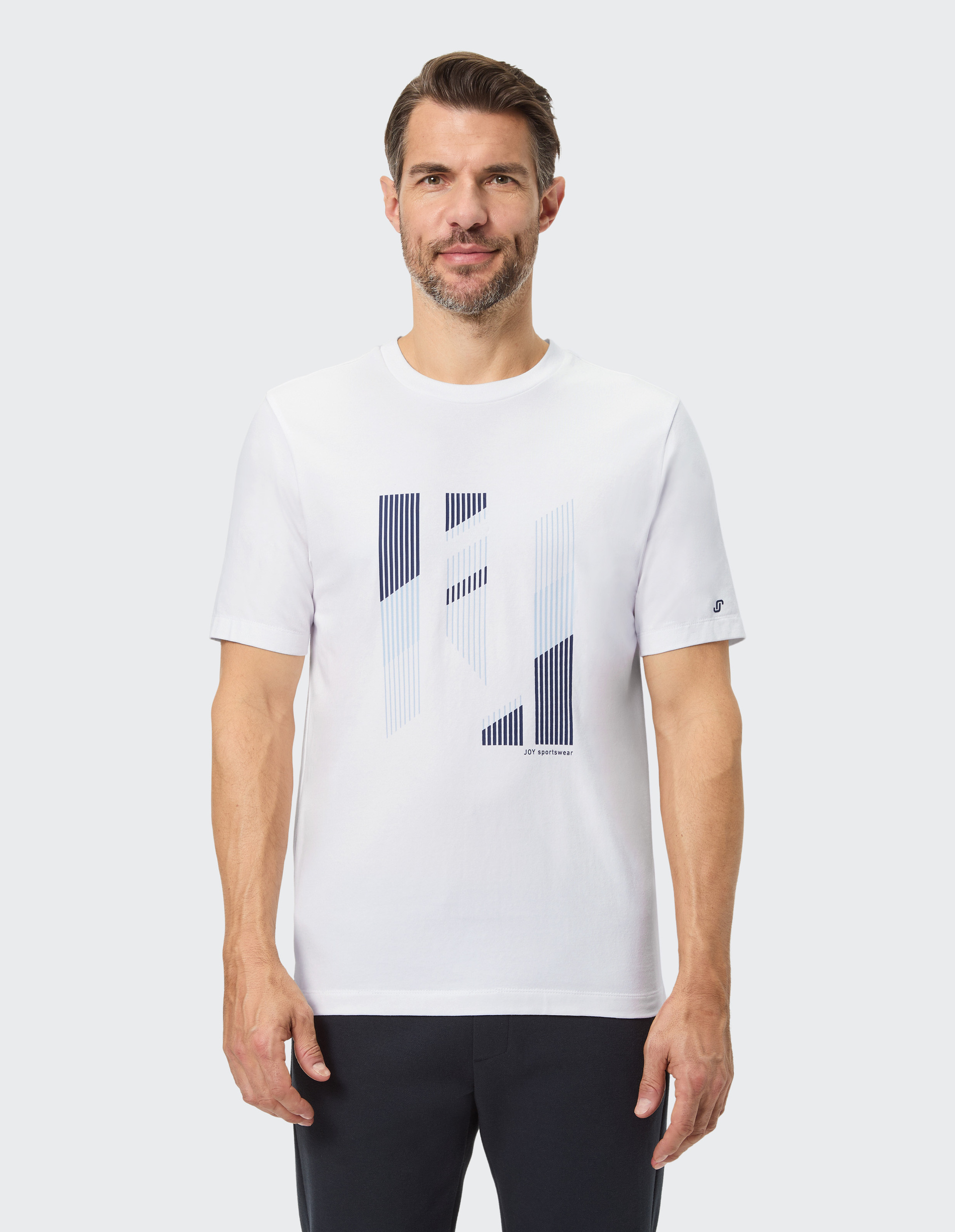 Ein Mann trägt ein weißes T-Shirt mit abstraktem Linienmuster vorne, kurze dunkle Haare, gepflegter Bart, und dunkle Hose.