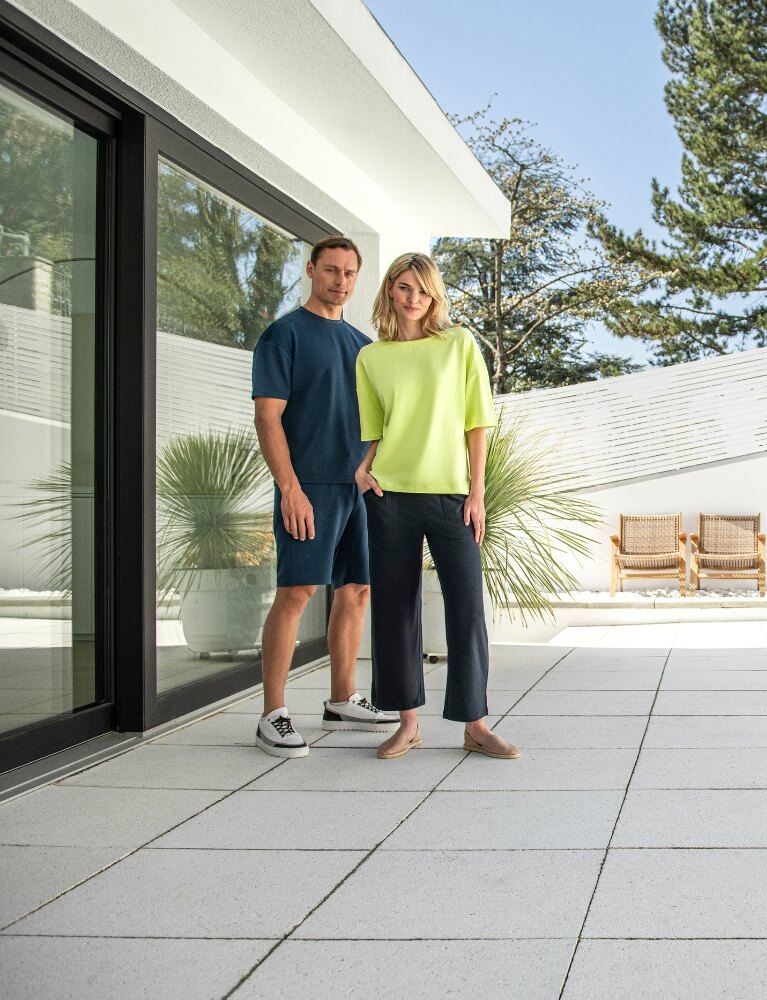 Mann in blauem Oberteil und blauer Hose von JOY sportswear steht neben Frau in gelbem Oberteil von JOY sportswear. Beide sehen frontal in die Kamera.