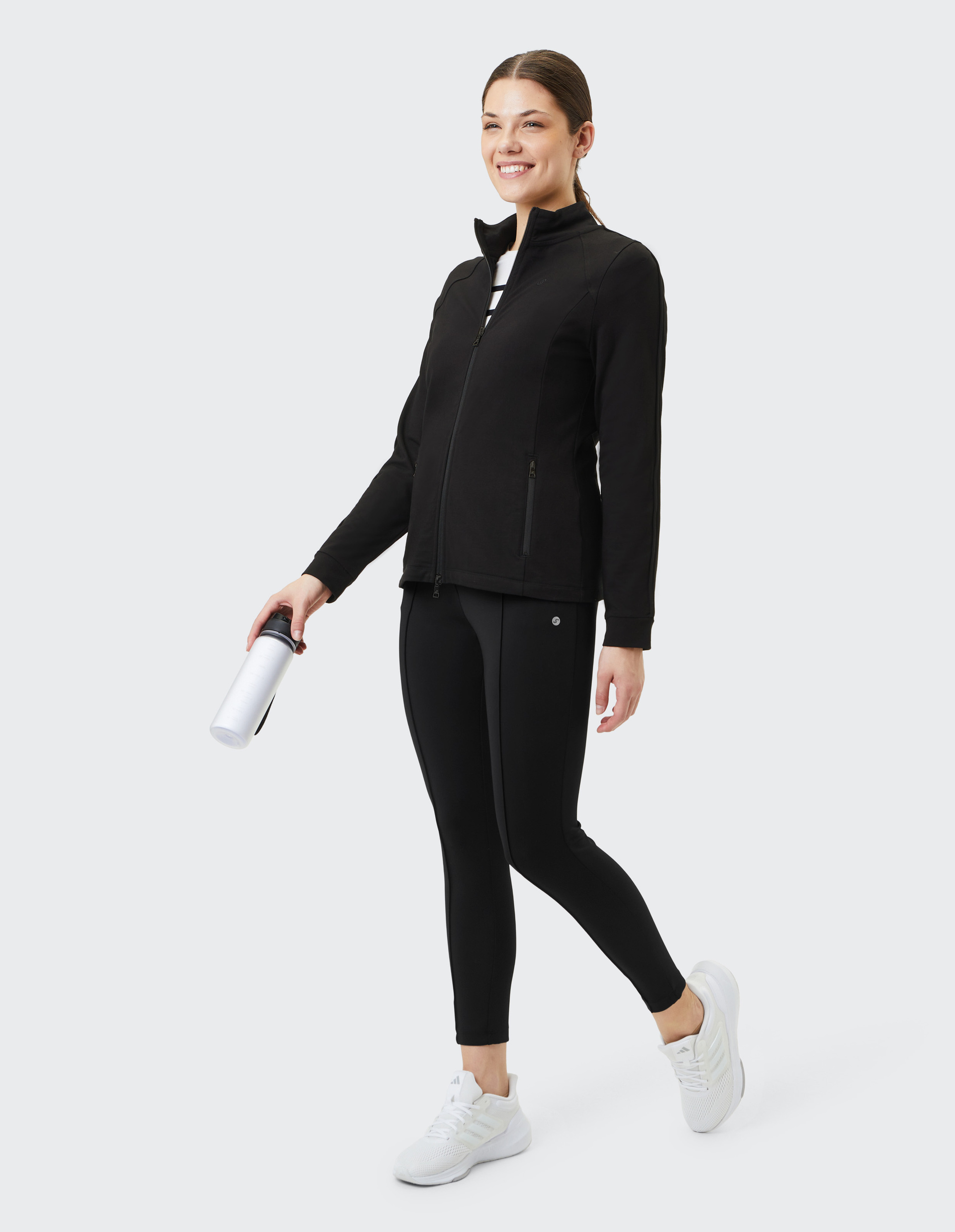 Frau in schwarzer Sportjacke und Leggings hält Wasserflasche und lächelt; trägt weiße Turnschuhe vor hellem Hintergrund.