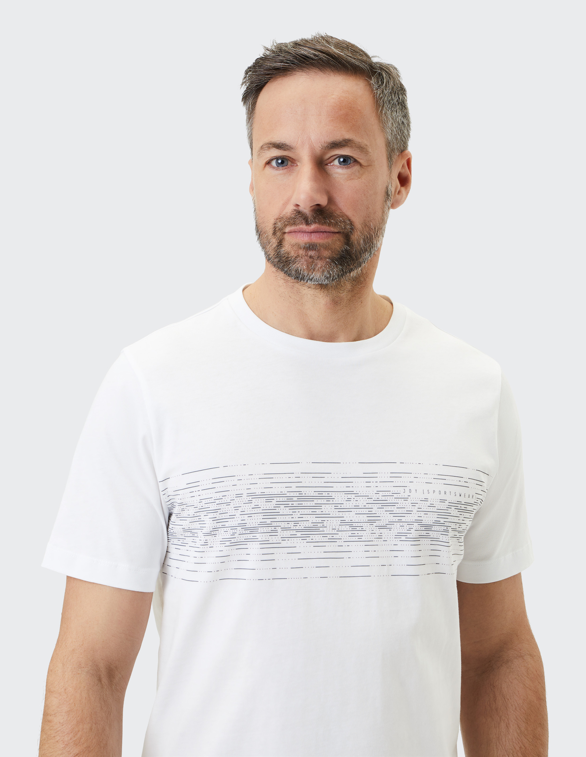 Ein Mann mit grauem Haar und Bart trägt ein weißes T-Shirt mit horizontalem, grauen Streifenmuster vor grauem Hintergrund.