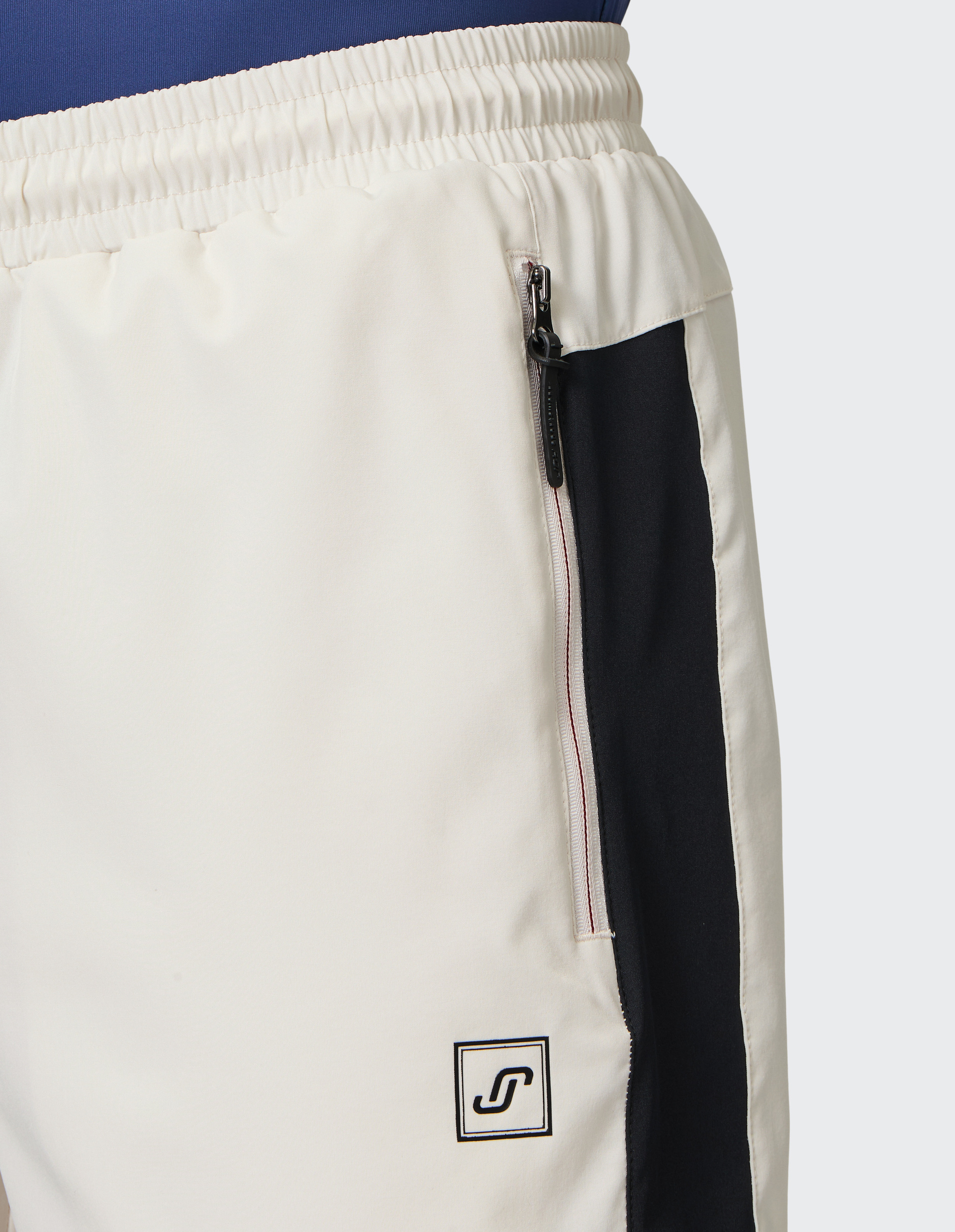 Nahaufnahme einer cremeweißen Jogginghose mit elastischem Bund, seitlichem schwarzen Streifen, Reißverschlusstasche und Logo.