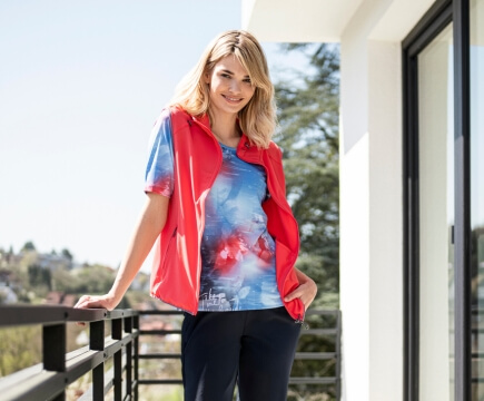 Frau mit roter Weste steht auf einem Balkon und stützt sich am Geländer ab.