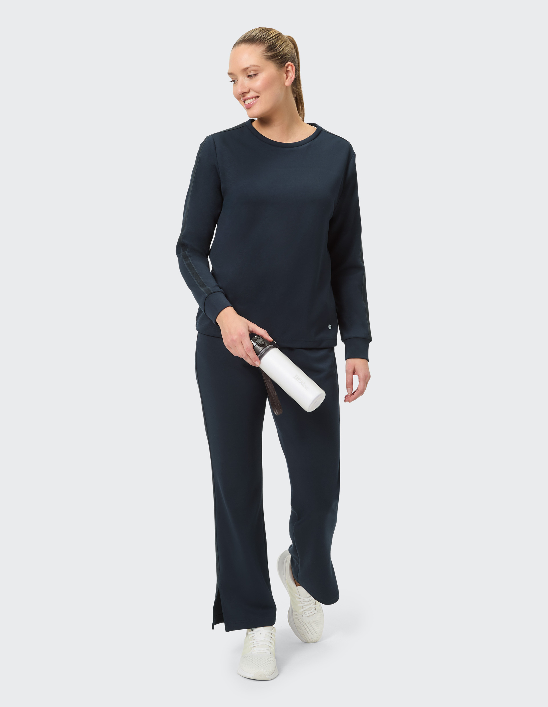 Frau trägt dunkelblauen Jogginganzug mit langen Ärmeln und weiten Hosen, weiße Sneaker, steht vor hellem Hintergrund.