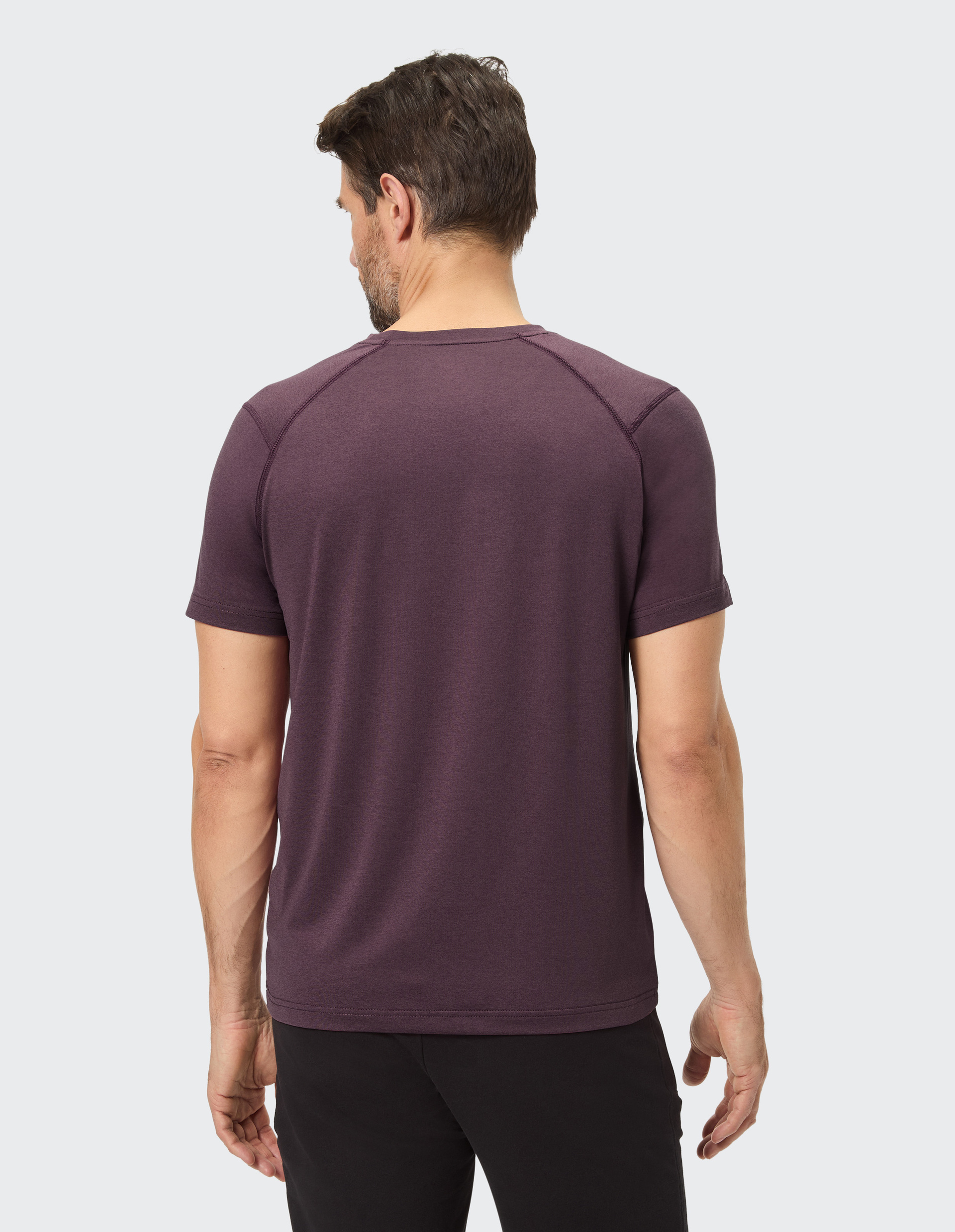 Ein Mann mit braunen Haaren trägt ein lila T-Shirt, fotografiert von hinten, vor hellem Hintergrund.