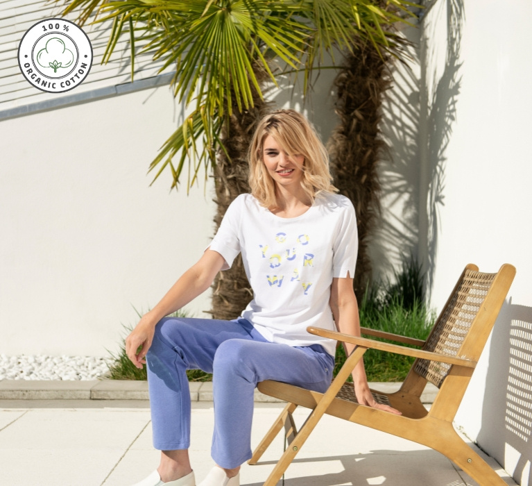 Frau sitzt in blauer Hose und weißem Shirt mit Druck von JOY sportswear an der Kante eines Liegestuhls. Links oben im Bild ist das Zeichen für Organic Cotton angebracht.