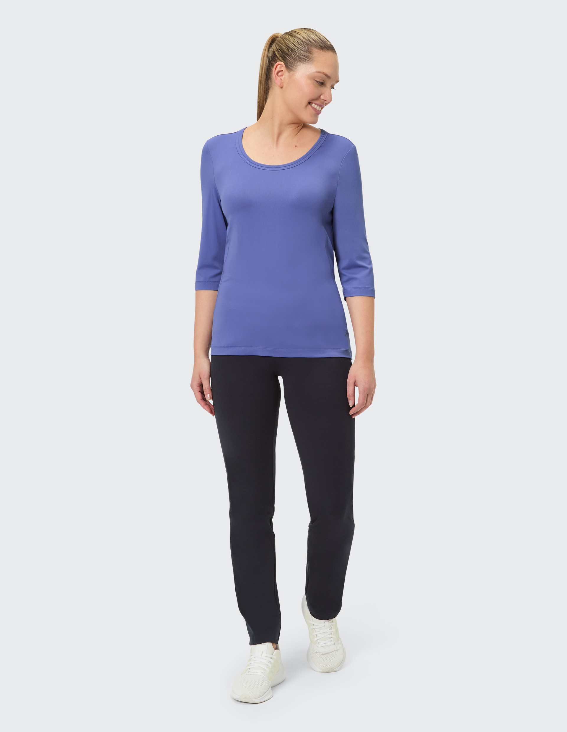 Frau mit blauer Bluse und schwarzer Hose, steht lächelnd, helle Sportschuhe, vor hellem Hintergrund.