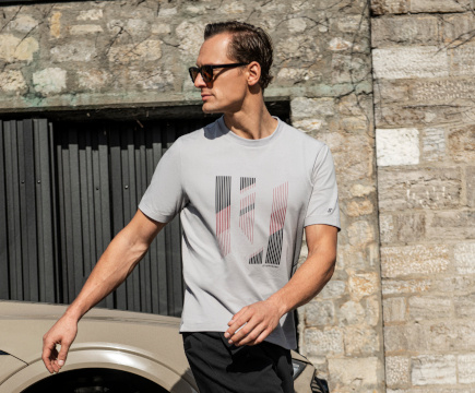 Mann mit Sonnenbrille in grauem T-Shirt mit grafischem Frontprint und schwarzer Hose der Marke JOY sportswear.