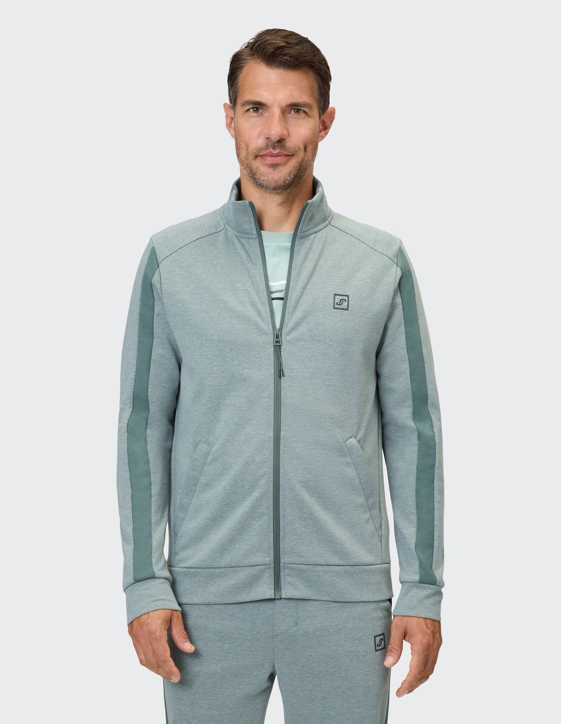 Ein Mann trägt eine hellgraue Trainingsjacke mit zwei vertikalen dunkelgrünen Streifen an den Ärmeln und einem Logo.