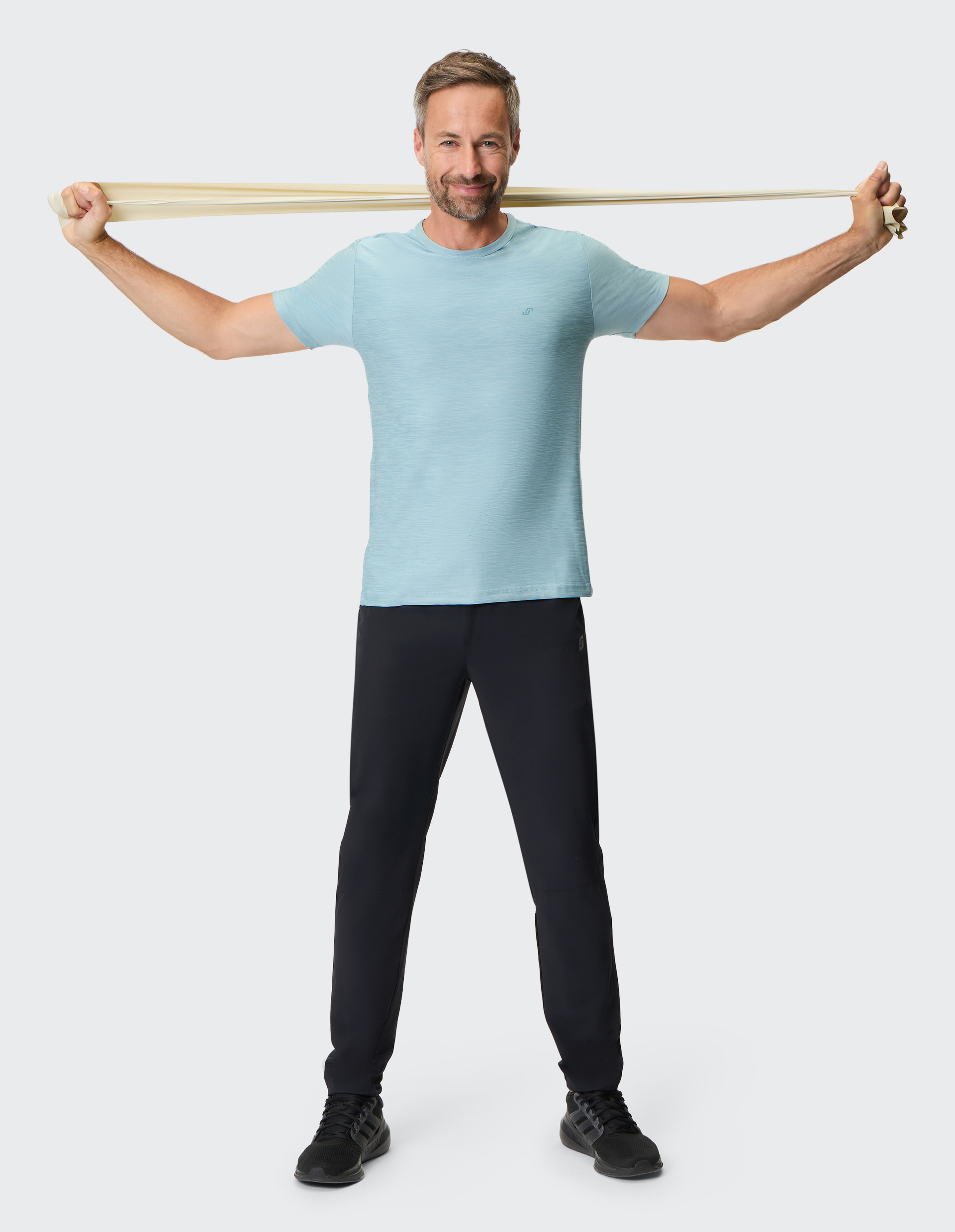 Ein lächelnder Mann in Sportkleidung hält ein Fitnessband hinter dem Rücken mit ausgestreckten Armen vor einem weißen Hintergrund.