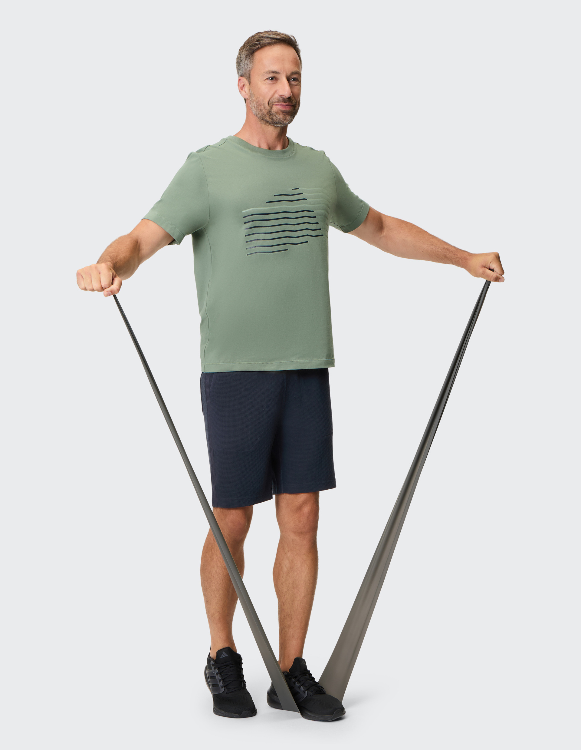 Mann in grünen T-Shirt und dunkelblauen Shorts trainiert mit einem Fitnessband, das unter seinen Füßen fixiert ist, Arme nach außen gestreckt.