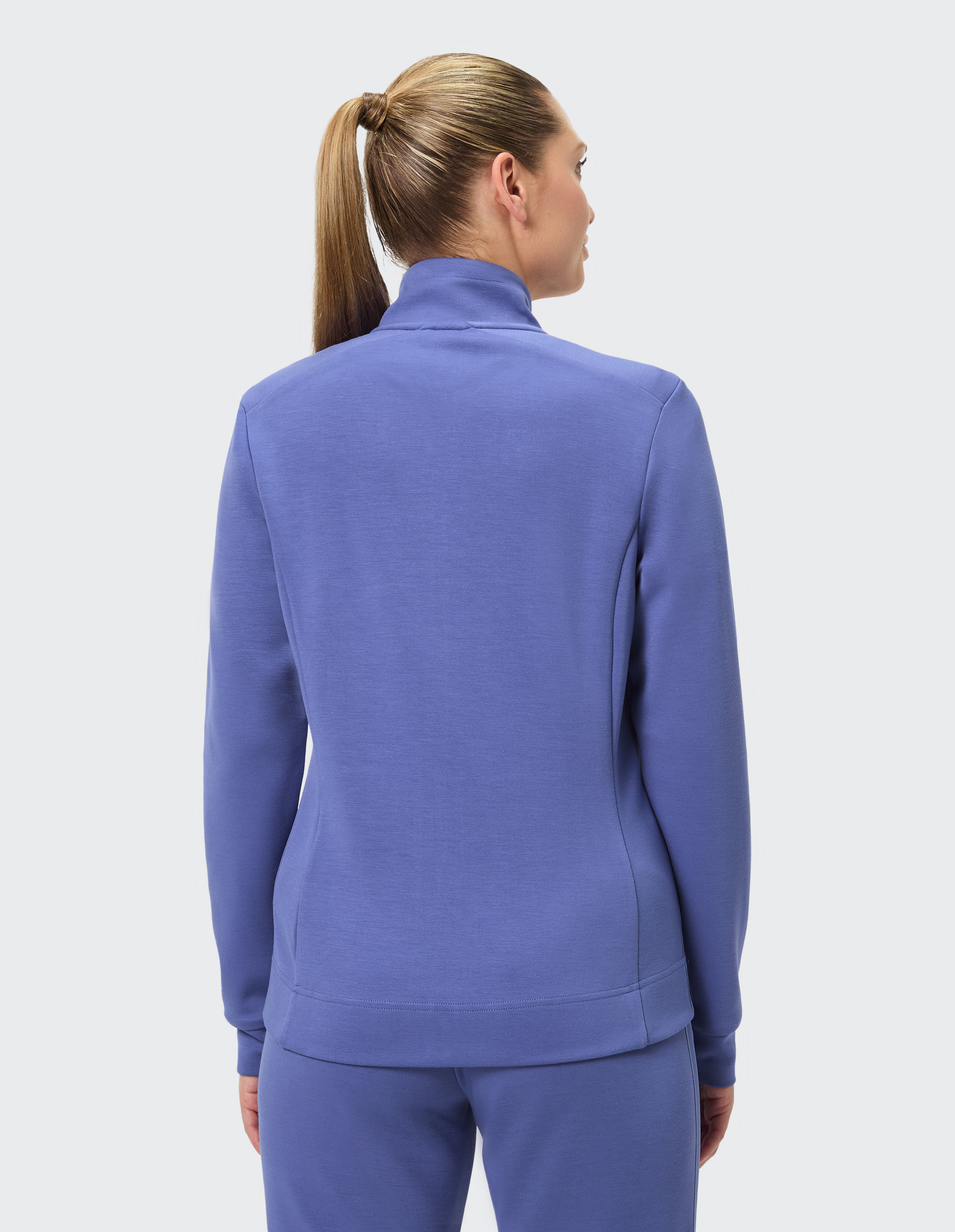 Frau in einem lila Trainingsanzug mit Pferdeschwanz von hinten, steht vor einem hellen Hintergrund.