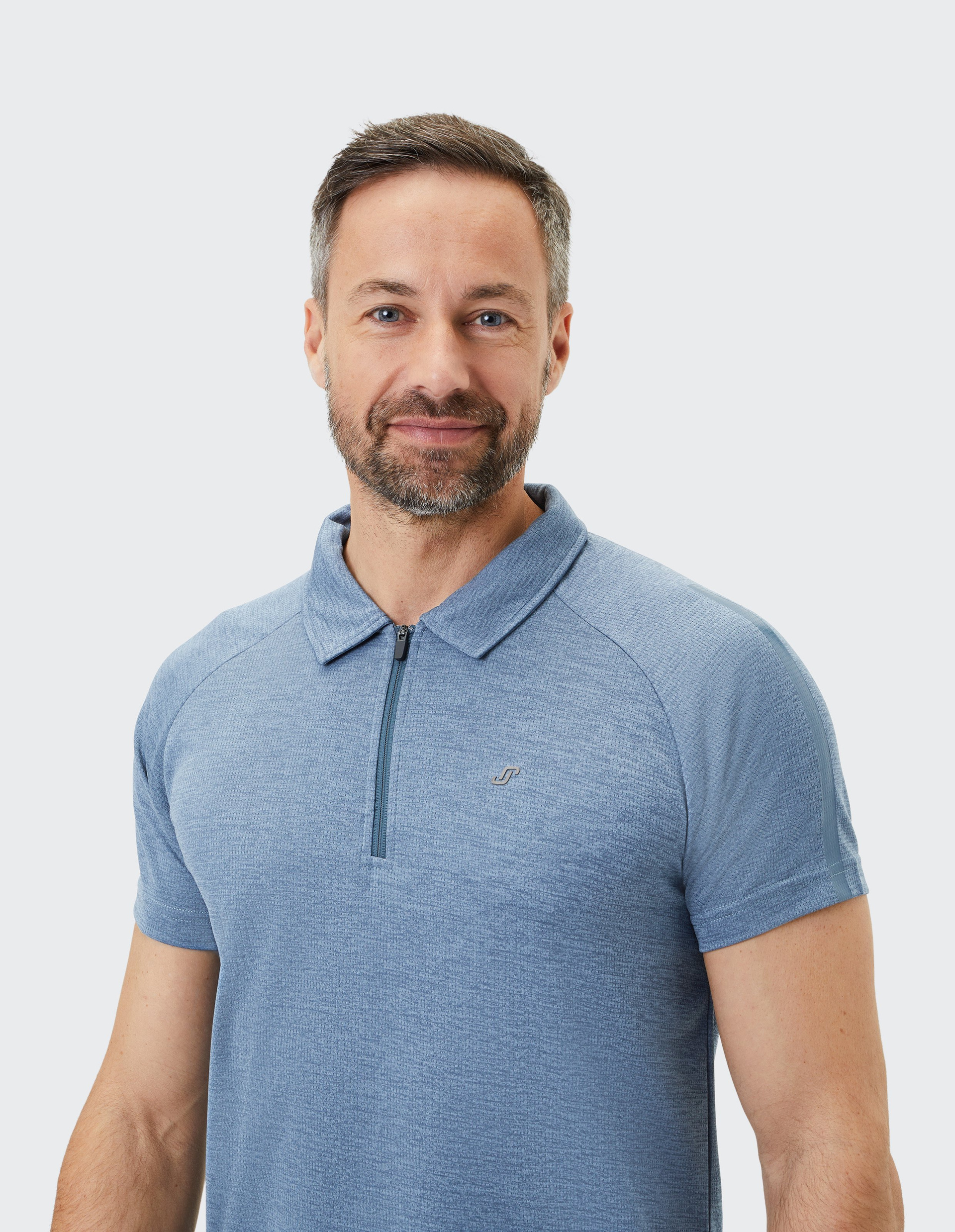 Ein lächelnder Mann mit Bart trägt ein hellblaues Polohemd mit Reißverschluss vor einem schlichten, weißen Hintergrund.