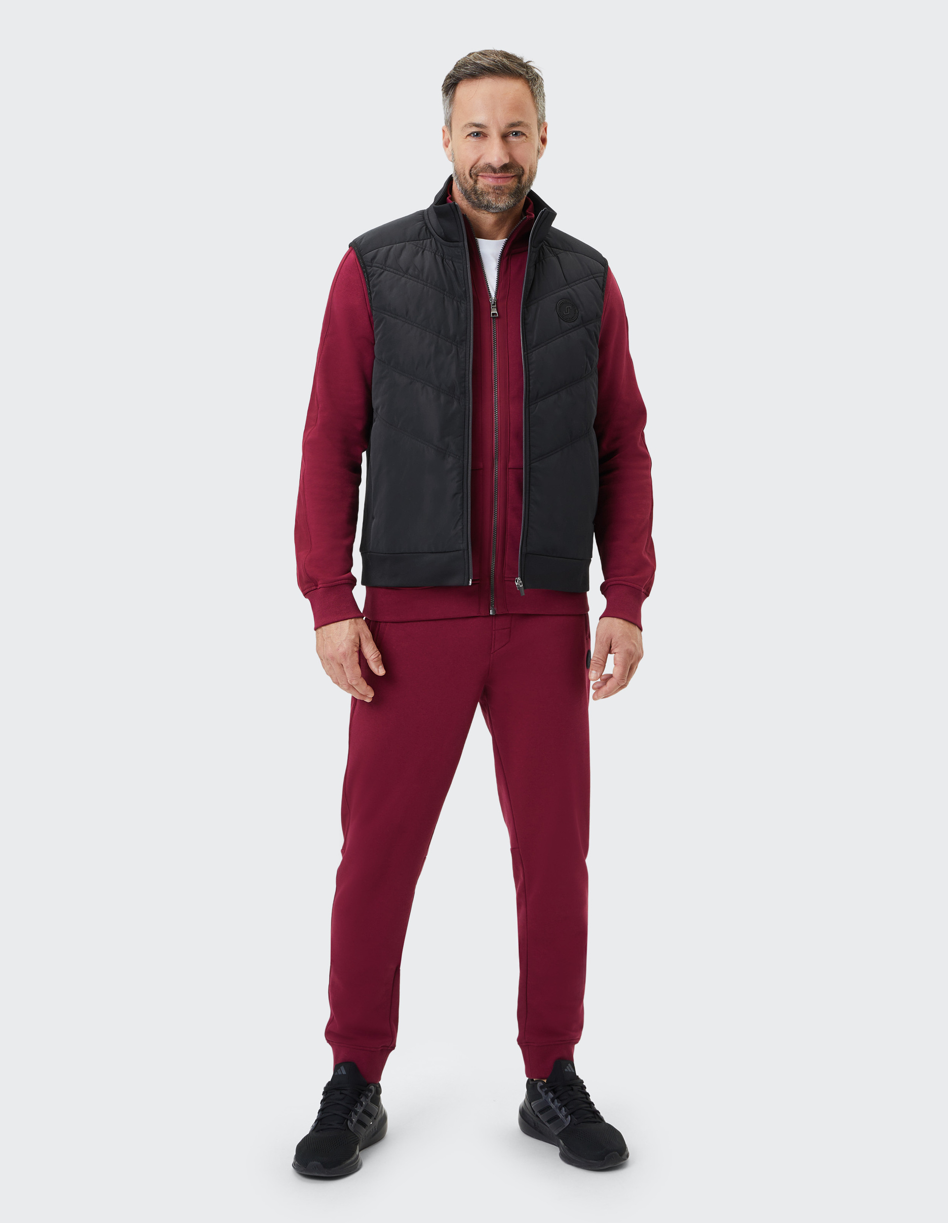 Ein Mann in einem weinroten Trainingsanzug mit schwarzer Weste steht lächelnd vor einem hellgrauen Hintergrund.