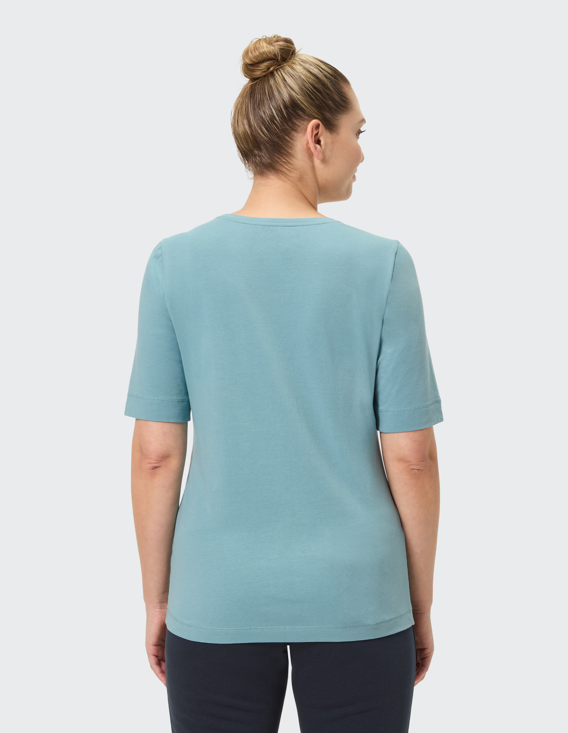 Frau mit Dutt trägt ein hellblaues T-Shirt und dunkelblaue Hose, Rückenansicht, vor hellem Hintergrund.