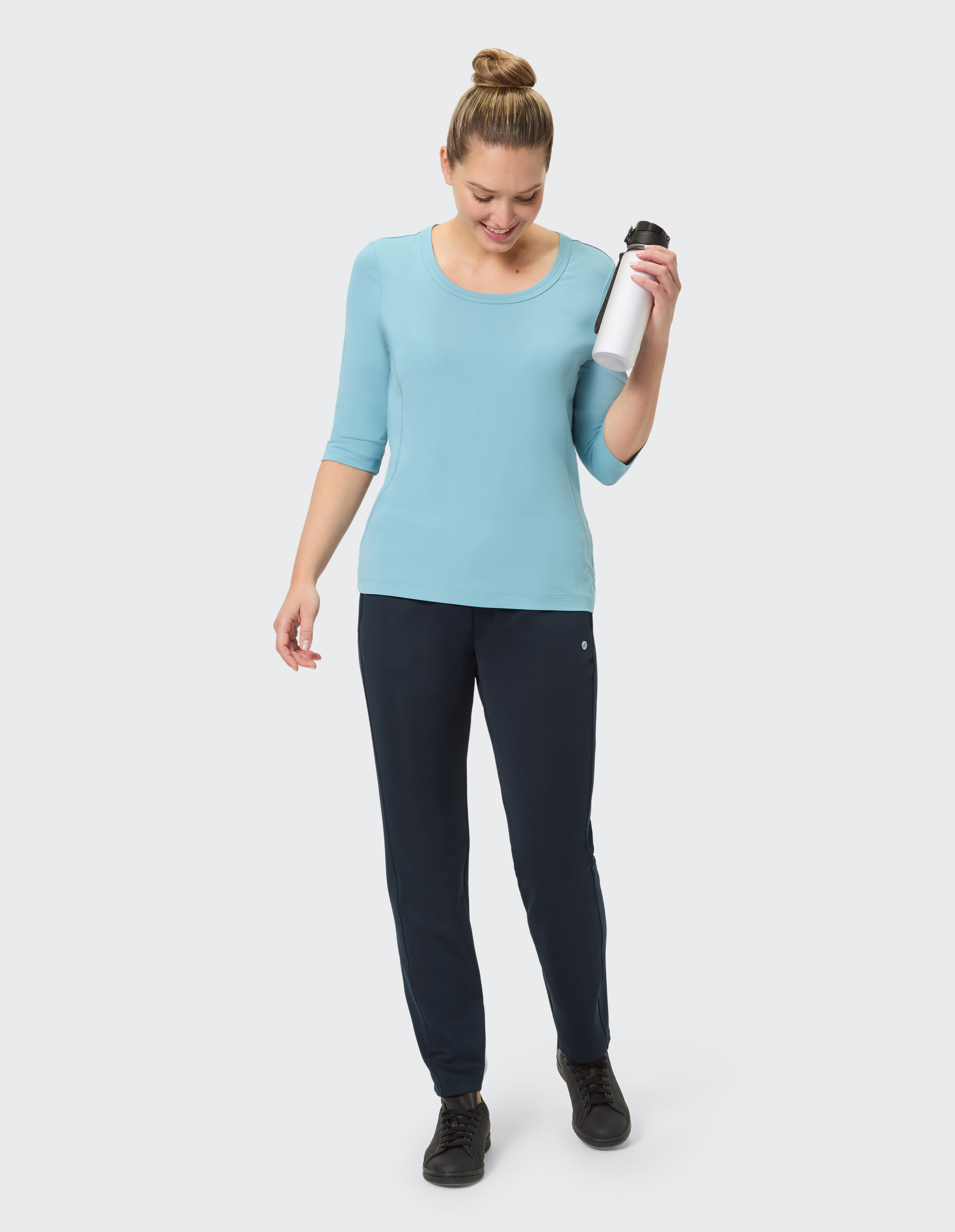 Frau in hellblauem Shirt und dunklen Hosen steht vor grauem Hintergrund, trägt schwarze Schuhe, lächelt sanft, Haare hochgesteckt.