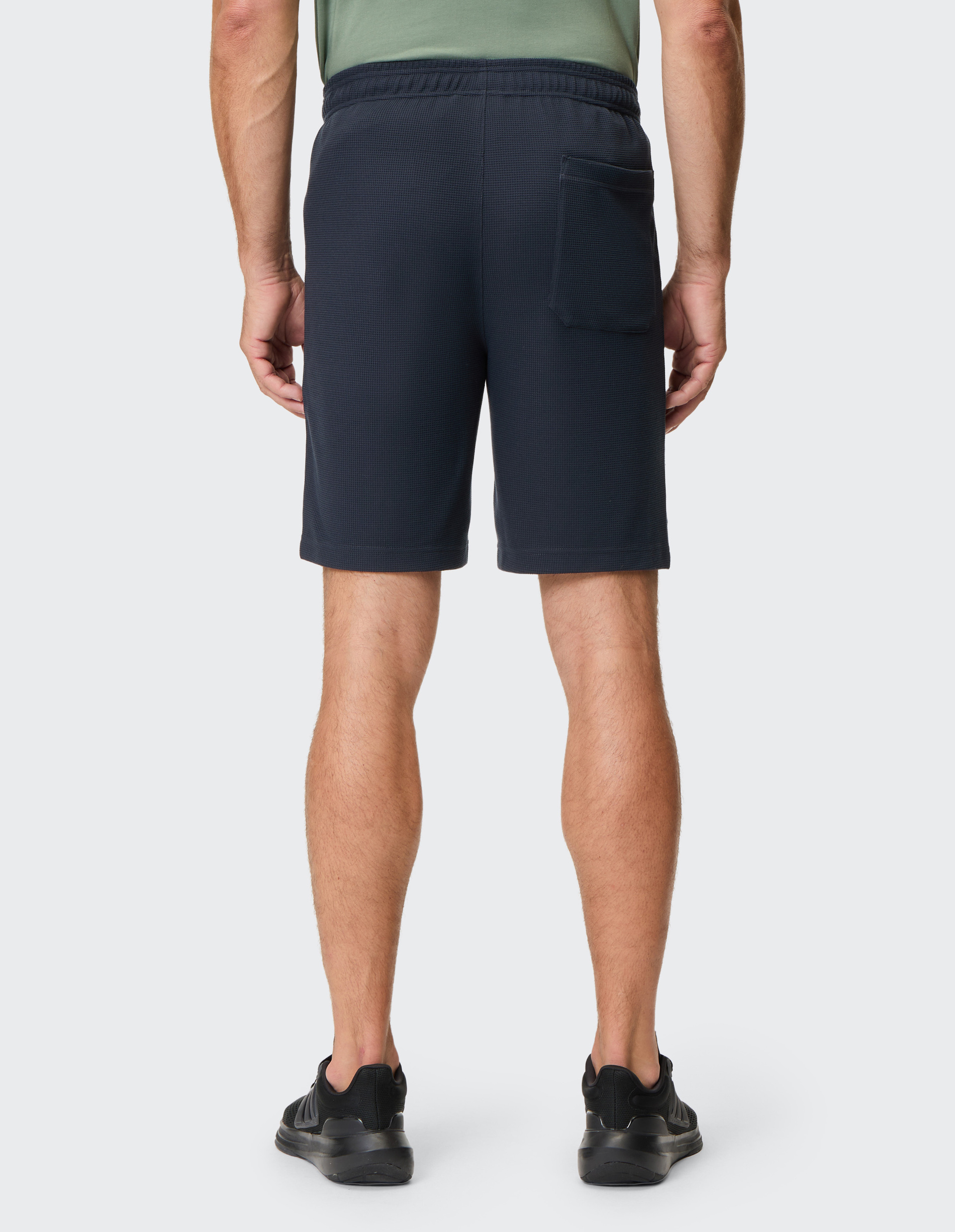 Ein Mann von hinten in dunkelblauen Shorts und schwarzem T-Shirt, trägt schwarze Sportschuhe auf neutralem Hintergrund.