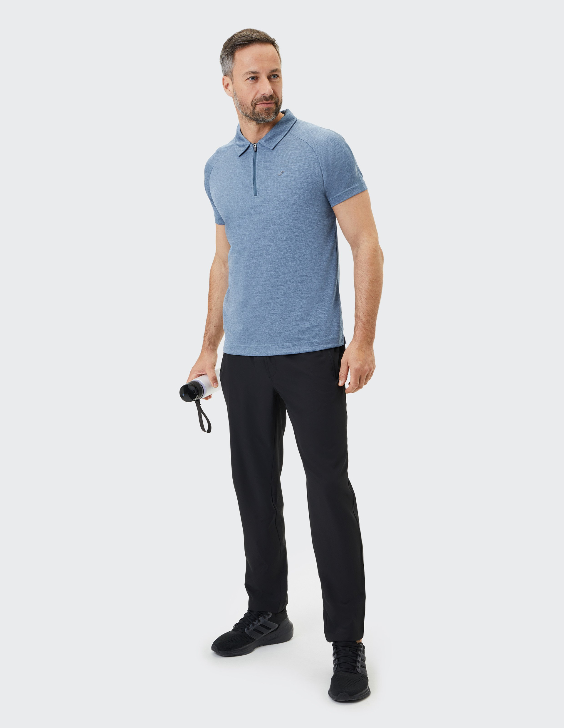 Mann in blauem Poloshirt und schwarzen Hosen hält eine Wasserflasche, steht selbstbewusst und schaut in die Ferne.