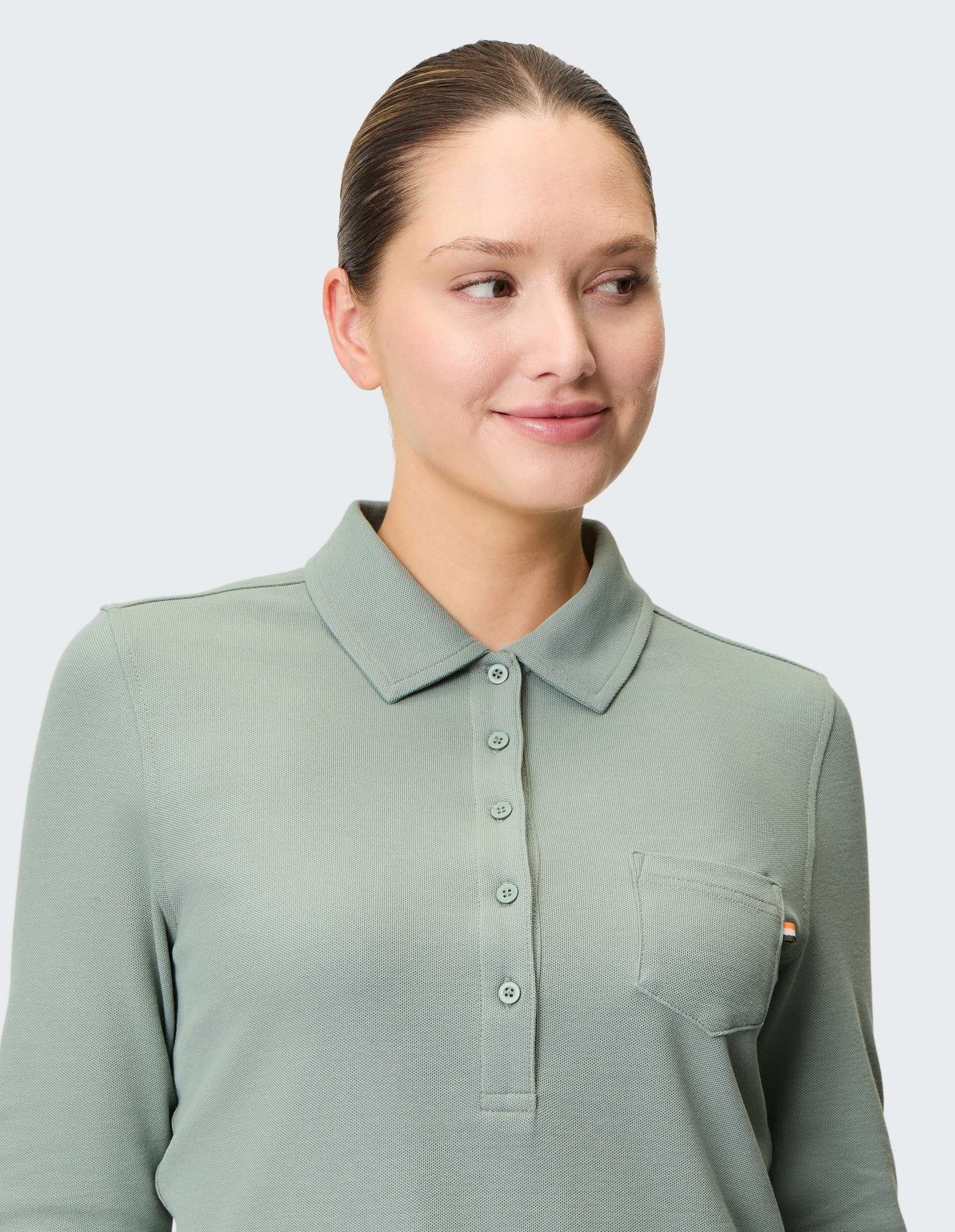 Eine Person mit zurückgebundenem Haar trägt ein grünes Polohemd mit Knopfleiste und Brusttasche, schaut lächelnd zur Seite.