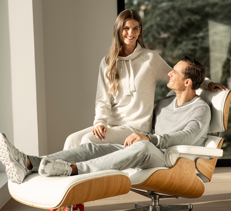 Mann in grauem Sweatshirt der Marke JOY sportswear sitz in einem weißen Sessel, auf der Lehne des Sessels sitzt eine Frau im beigefarbenen Jogginganzug.
