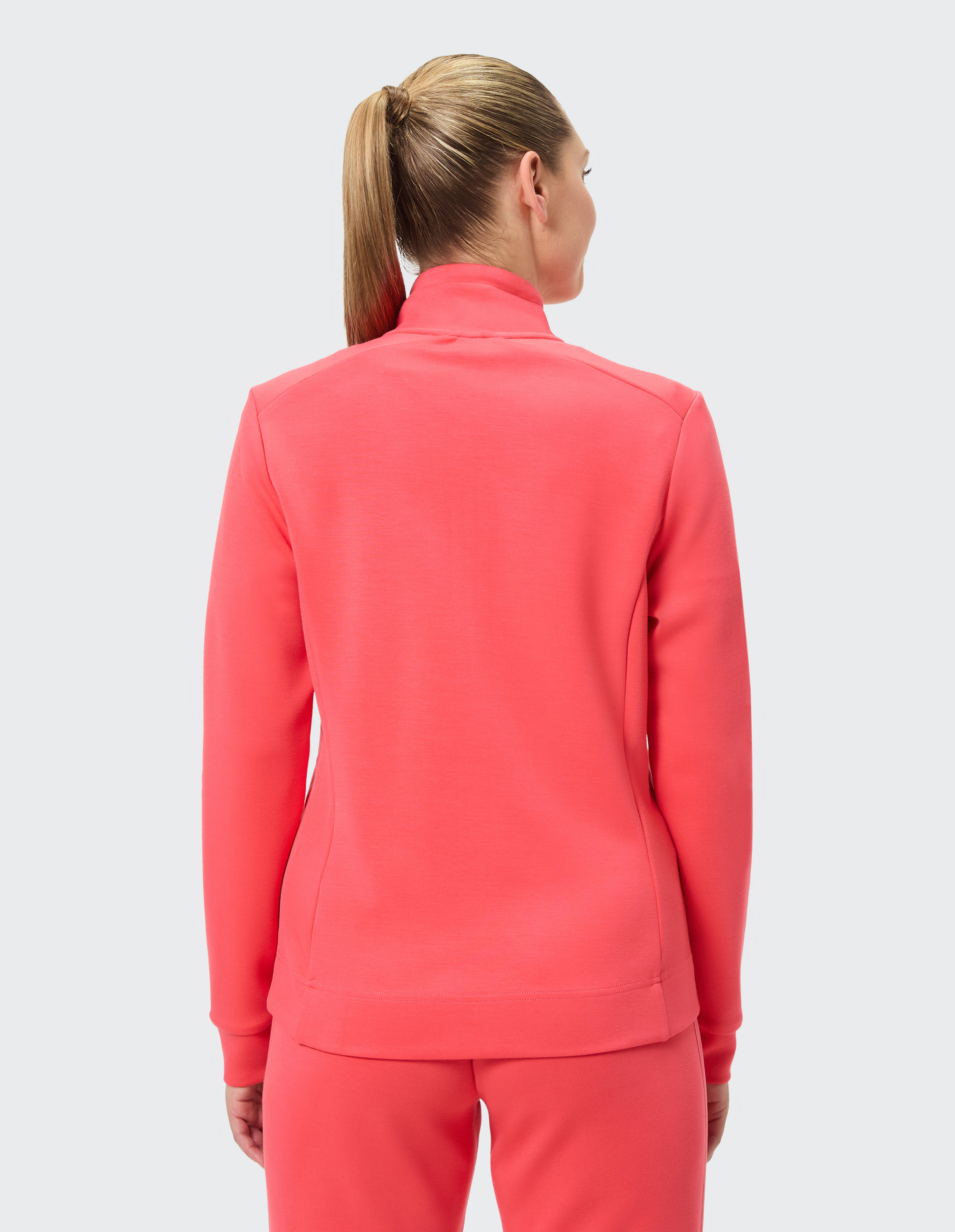 Eine Frau mit Pferdeschwanz trägt eine korallfarbene Trainingsjacke und Hose, von hinten vor hellem Hintergrund gesehen.