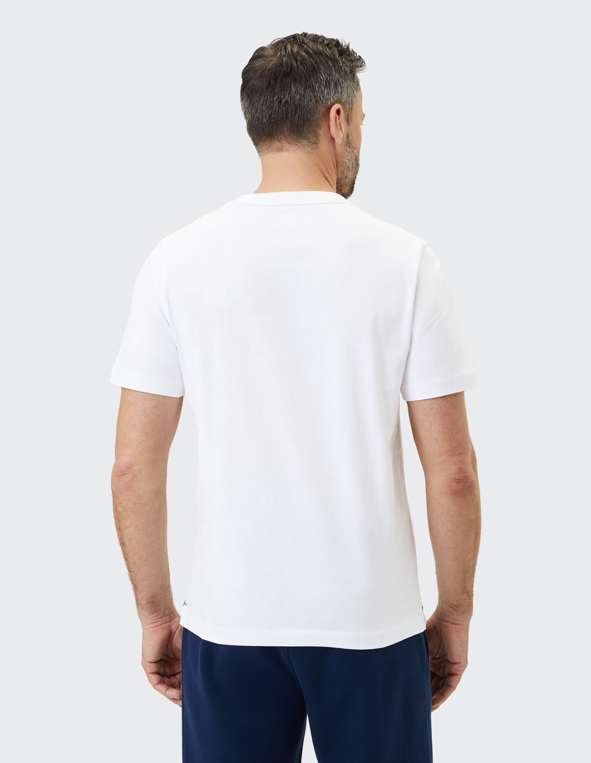 Mann mit kurzen Haaren trägt ein weißes T-Shirt und dunkelblaue Hose, steht mit dem Rücken zur Kamera vor grauem Hintergrund.