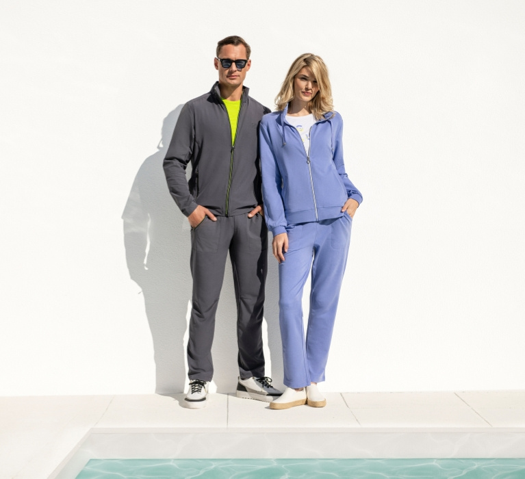 Frau in blauem Anzug steht neben Mann in grauem Anzug der Marke JOY sportswear. Vor den beiden ist die Ecke eines Pools zu sehen.