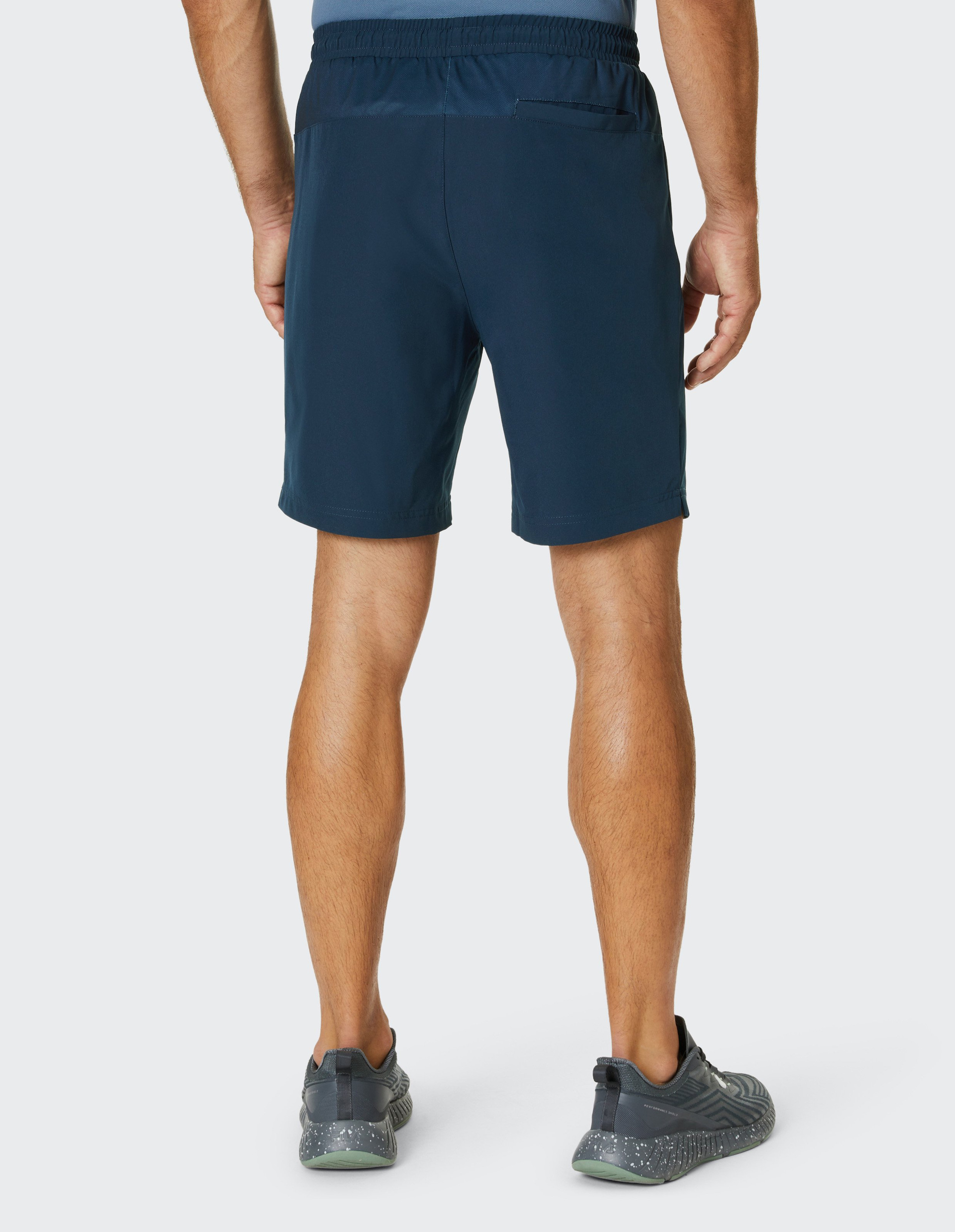 Ein Mann in dunkelblauen Sportshorts und grauen Turnschuhen steht mit dem Rücken zur Kamera auf einem weißen Hintergrund.