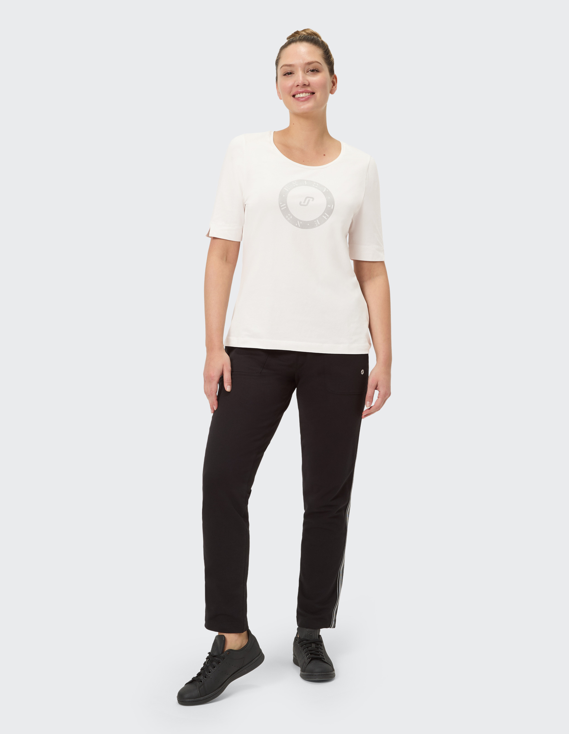 Frau mit Dutt trägt ein weißes Shirt mit grauem Logo und schwarze Hose mit Streifen, steht vor grauem Hintergrund.