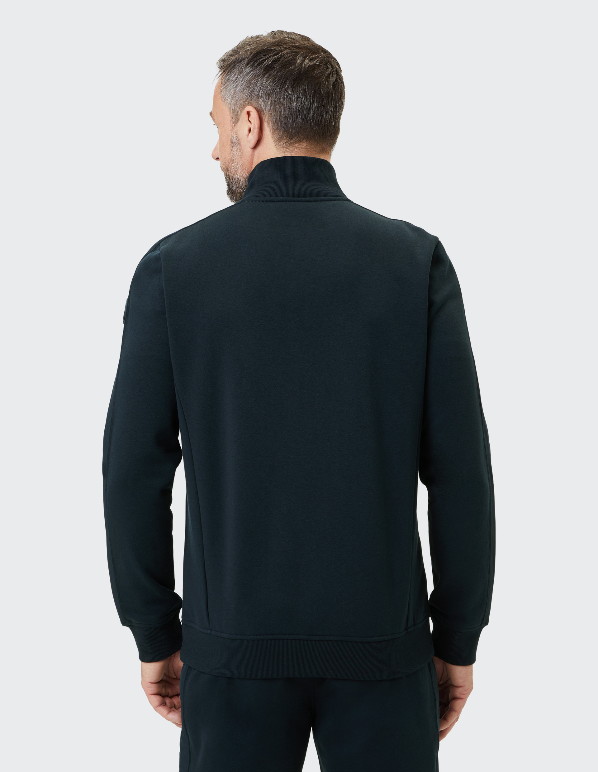 Mann von hinten in dunkelblauem Pullover mit Stehkragen, kurzgeschnittenem Haar, steht vor einem weißen Hintergrund.