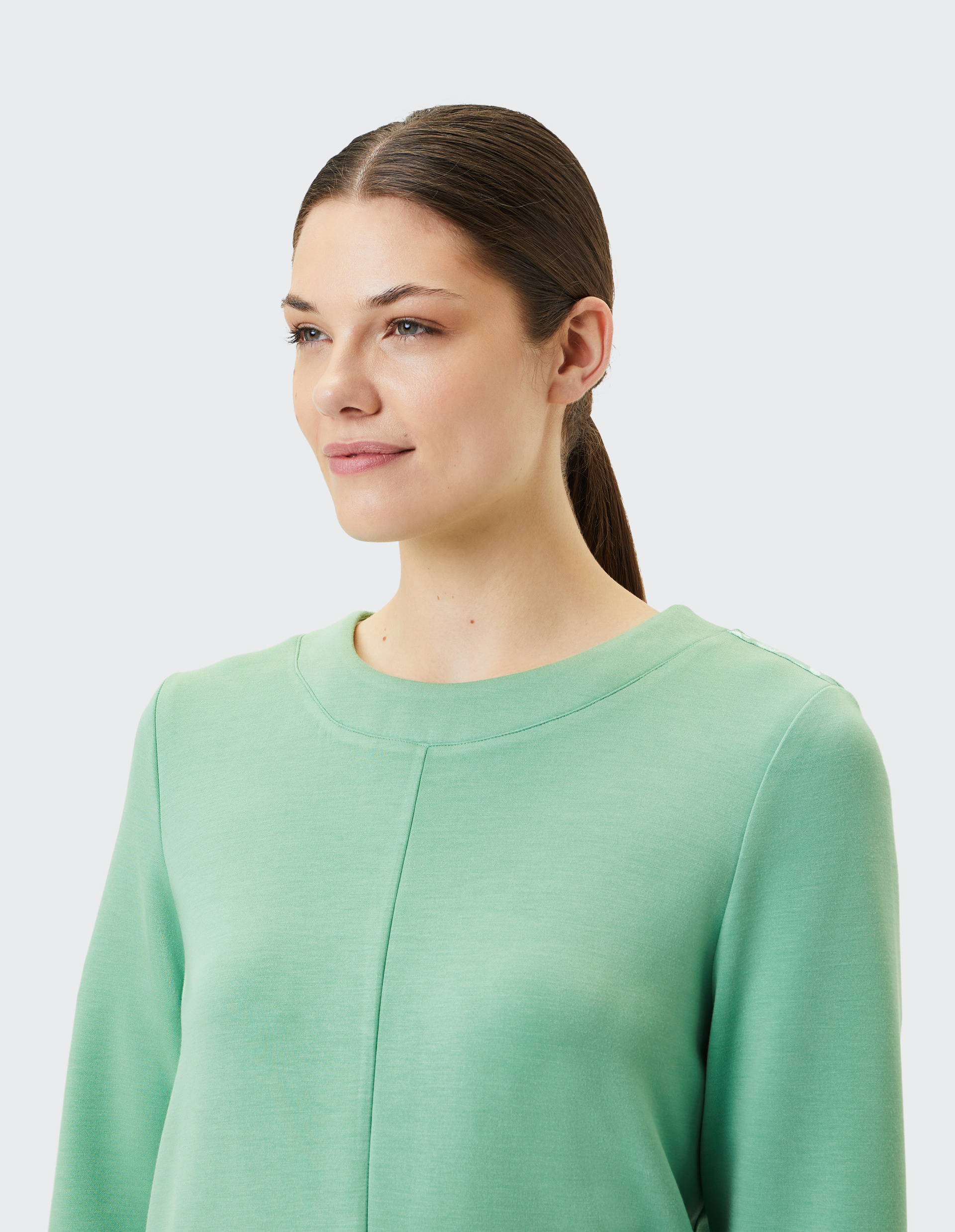 Junge Frau mit glattem Haar trägt einen mintgrünen Pullover und schaut lächelnd zur Seite, vor einem hellen Hintergrund.