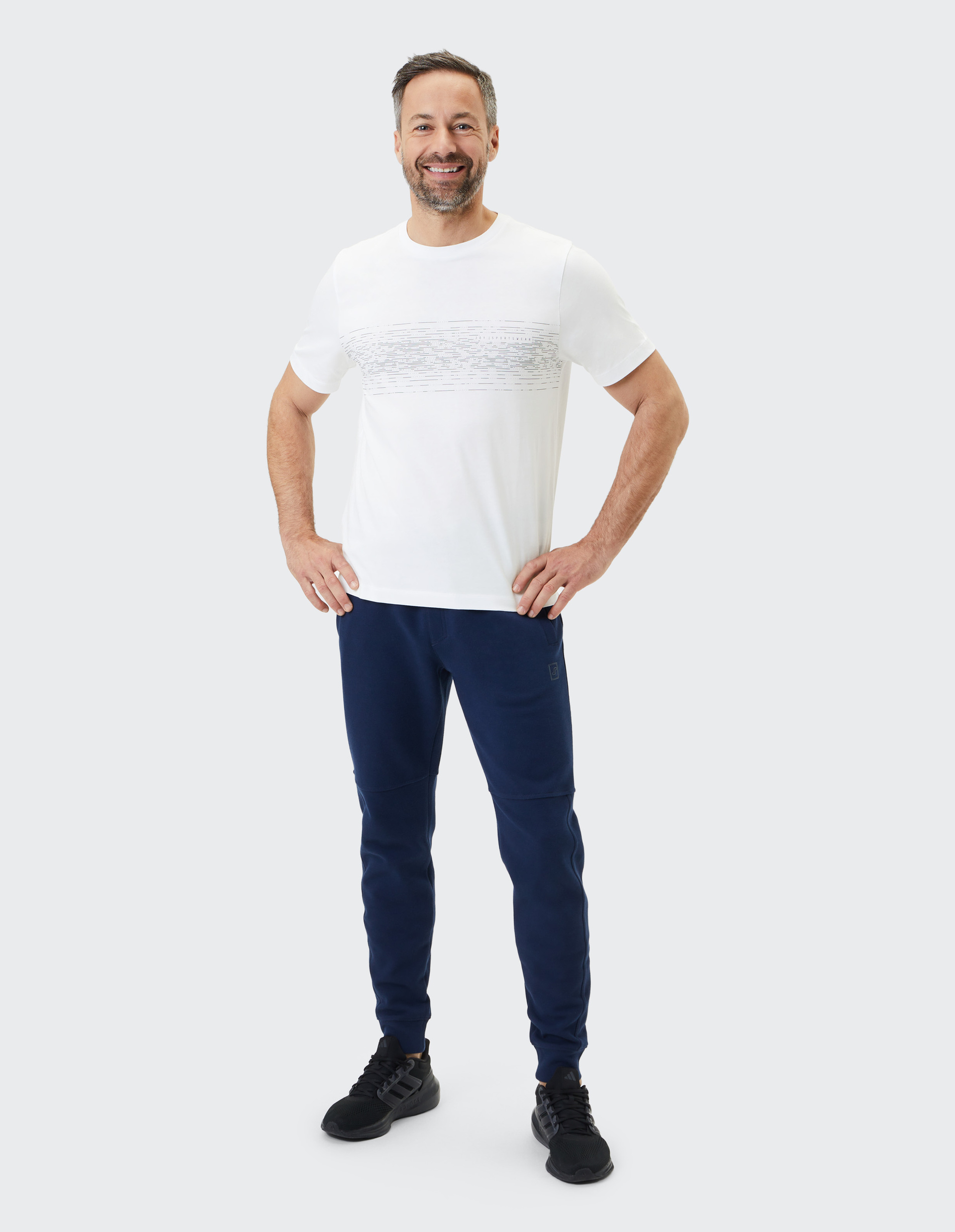 Ein lächelnder Mann steht in einem weißen T-Shirt und dunkelblauer Jogginghose vor neutralem Hintergrund, Hände in die Hüften gestemmt.