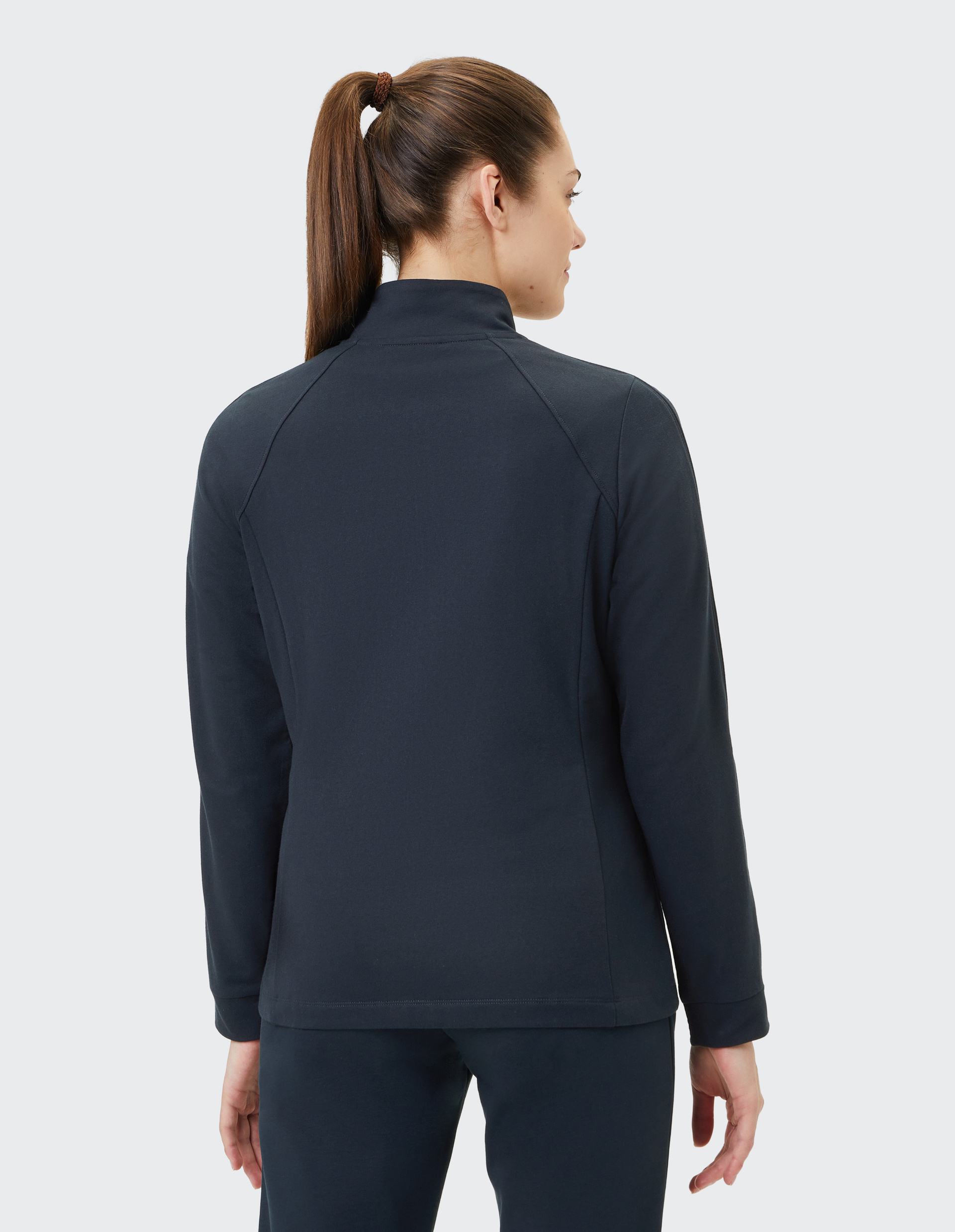 Frau mit Pferdeschwanz trägt eine dunkelblaue Jacke und blickt nach rechts; sie steht vor einem hellgrauen Hintergrund.