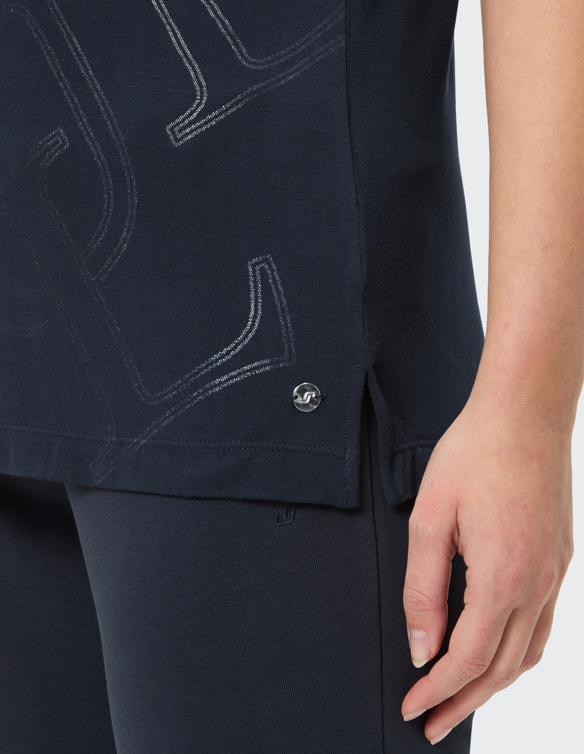 Ein dunkelblaues Oberteil mit silbernem Schriftzug und seitlichem Metallknopf, getragen von einer Person mit der Hand am Oberschenkel.