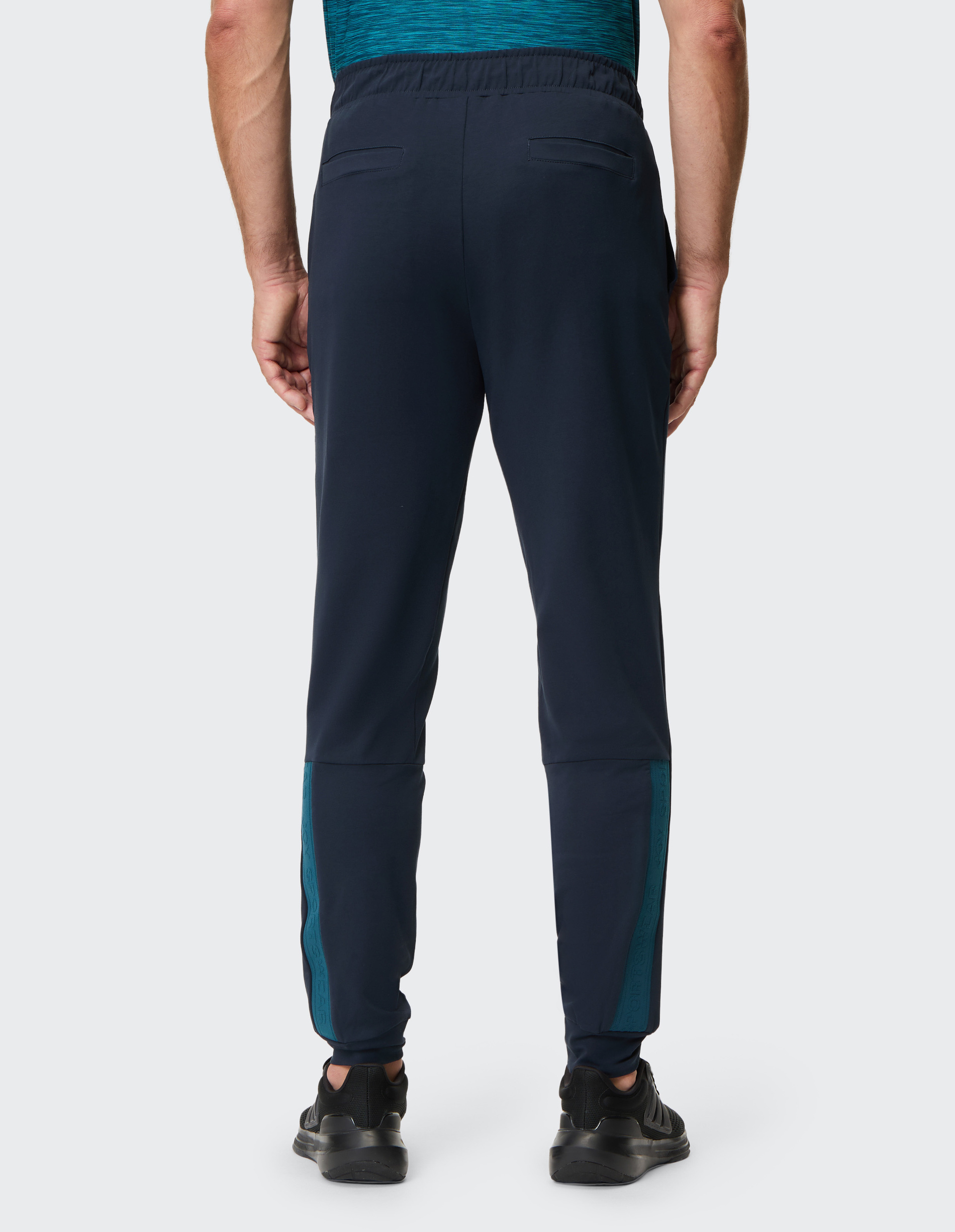 Der Mann trägt eine dunkelblaue Hose mit türkisen Streifen und schwarzen Schuhen, Rücken zur Kamera, auf hellem Hintergrund.