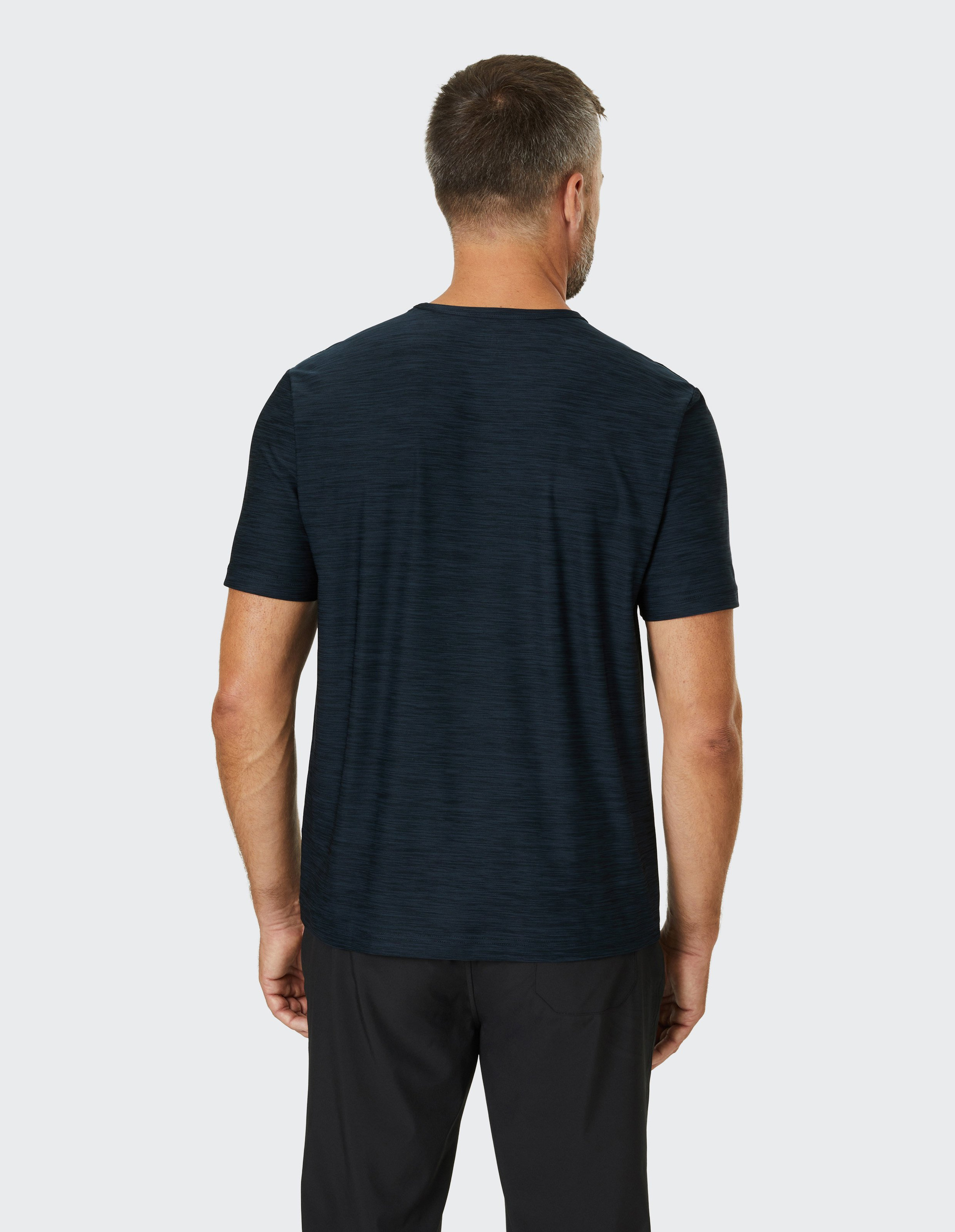 Ein Mann in schwarzem T-Shirt und Hose steht mit dem Rücken zur Kamera vor hellem Hintergrund.